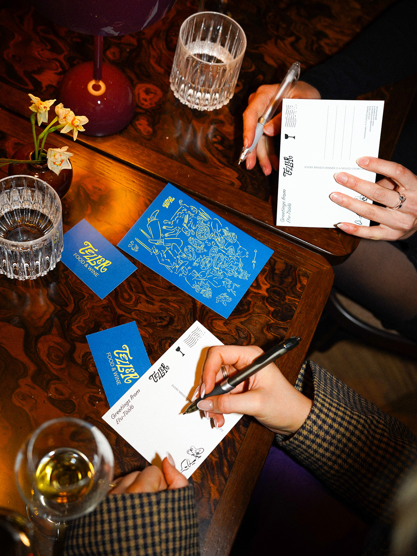 Two people writing on Teller Food & Wine postcards at a wooden table with blue branded cards and glassware.