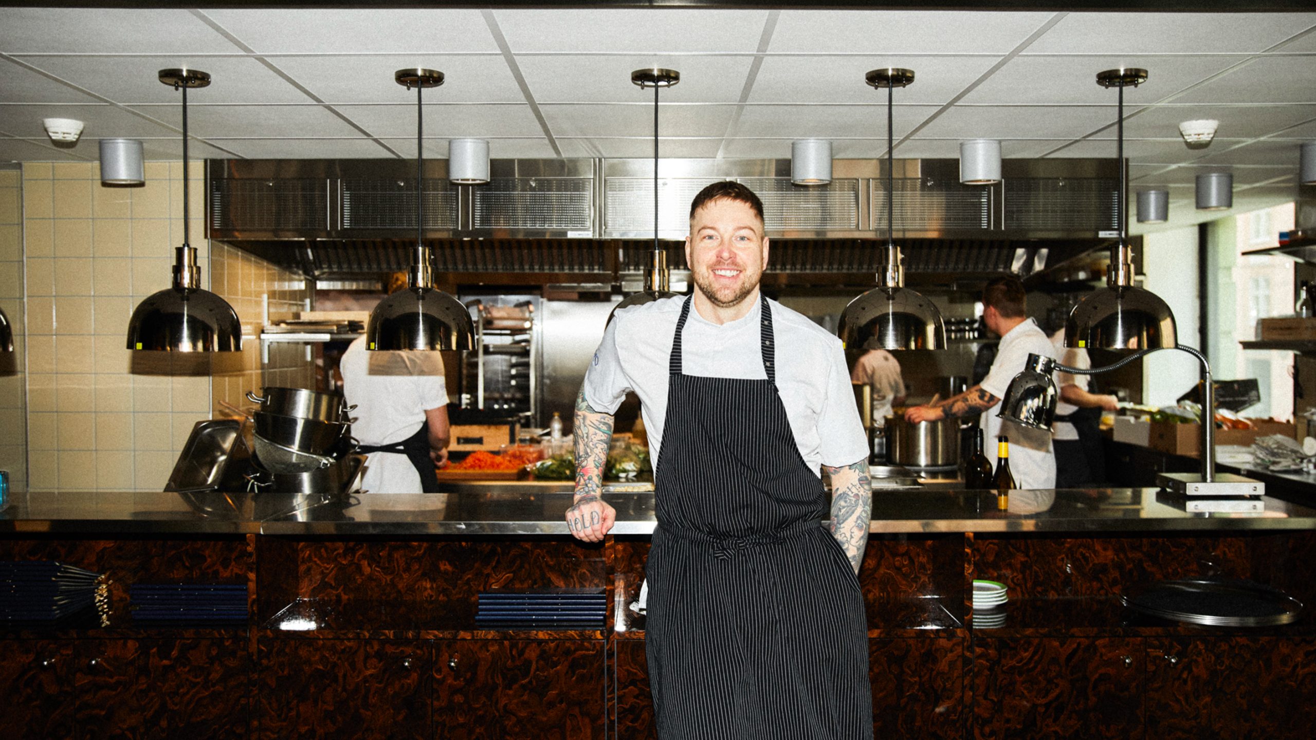 Chef in striped apron smiling at Teller restaurant's open kitchen counter, staff working in background.