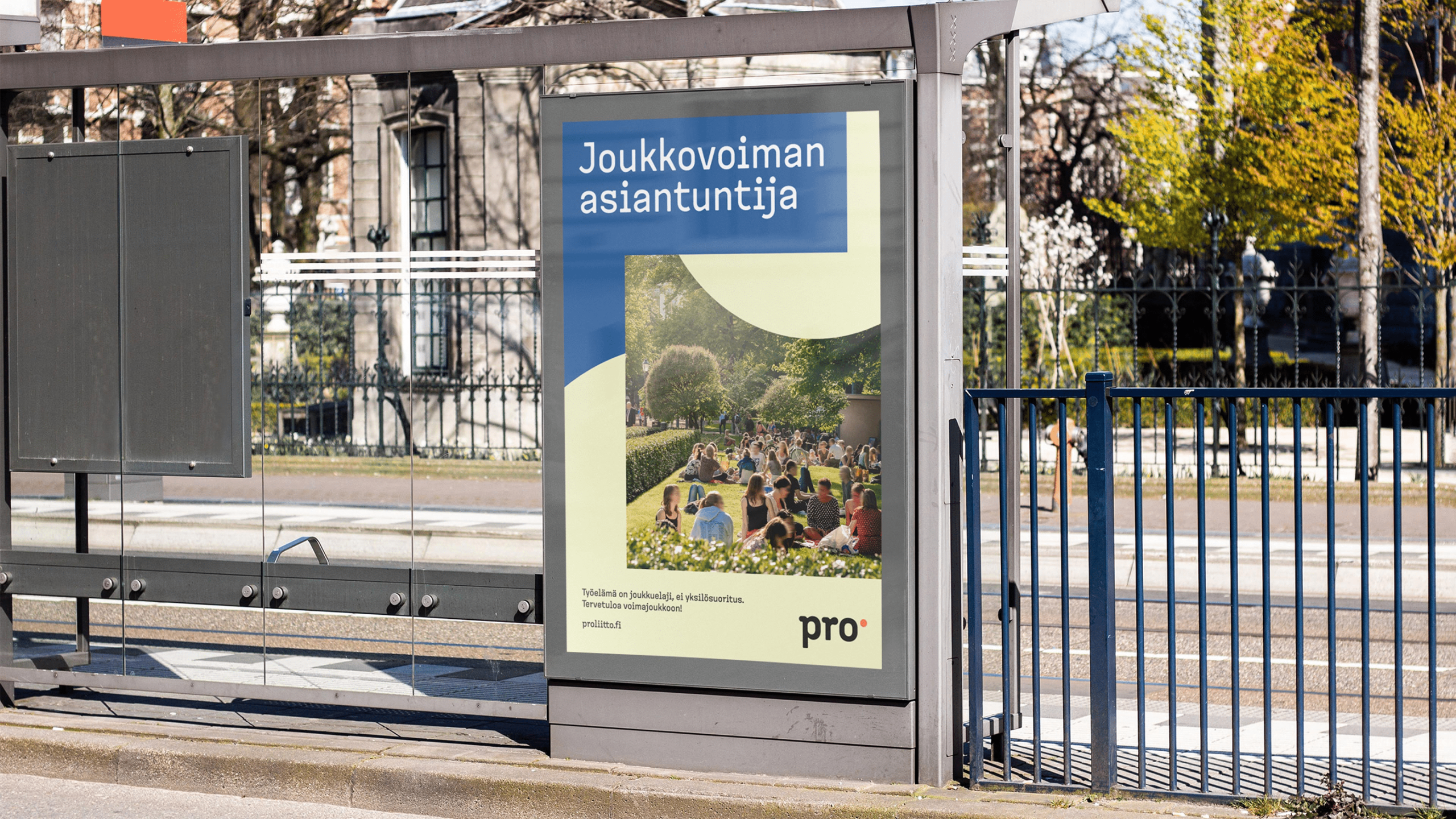 Pro liitto bus stop poster reading 'Joukkovoiman asiantuntija' with a crowd on grass, in blue and cream tones.