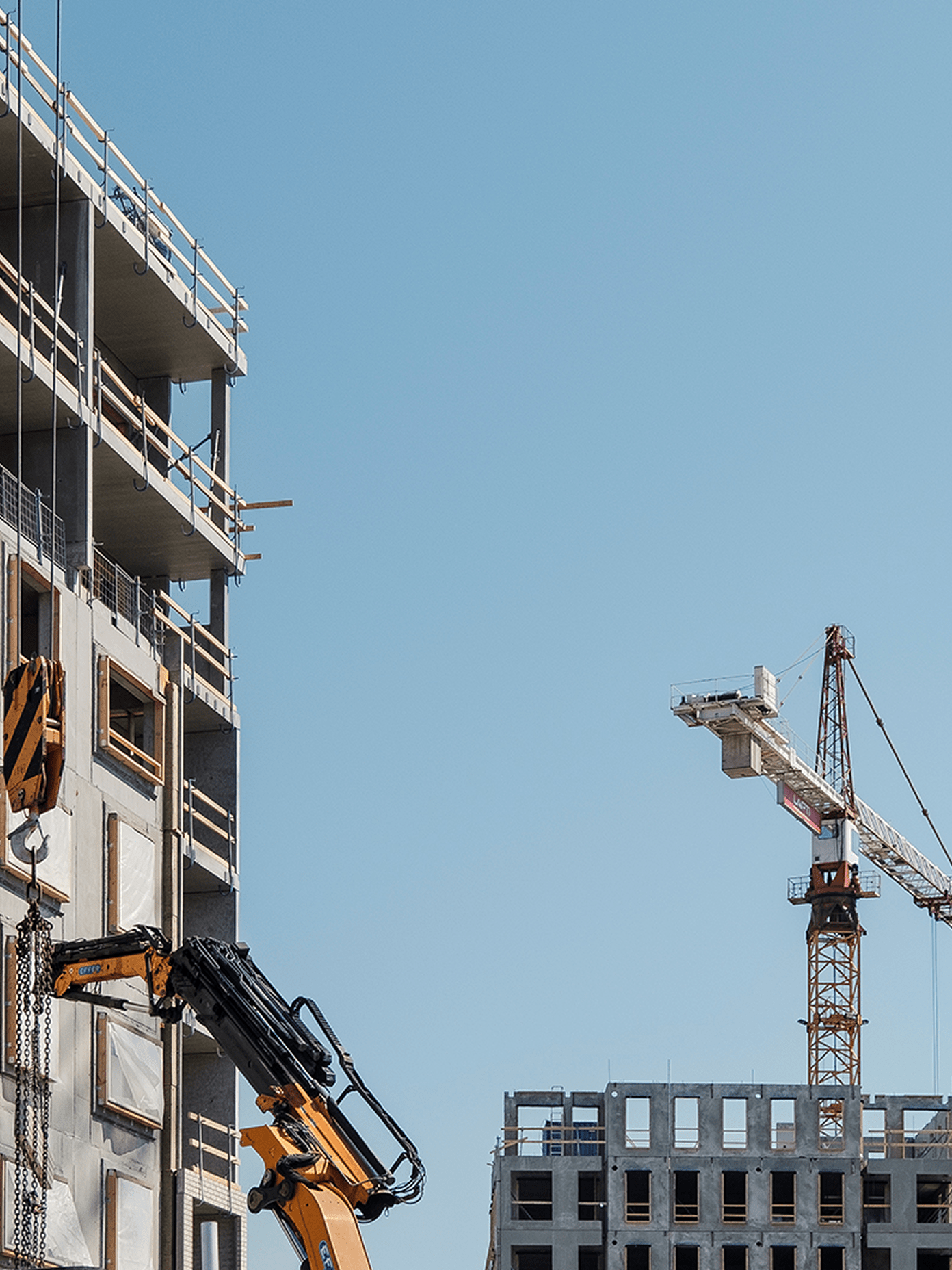 Construction site with concrete building frames, a yellow crane arm, and a tower crane against a clear blue sky.