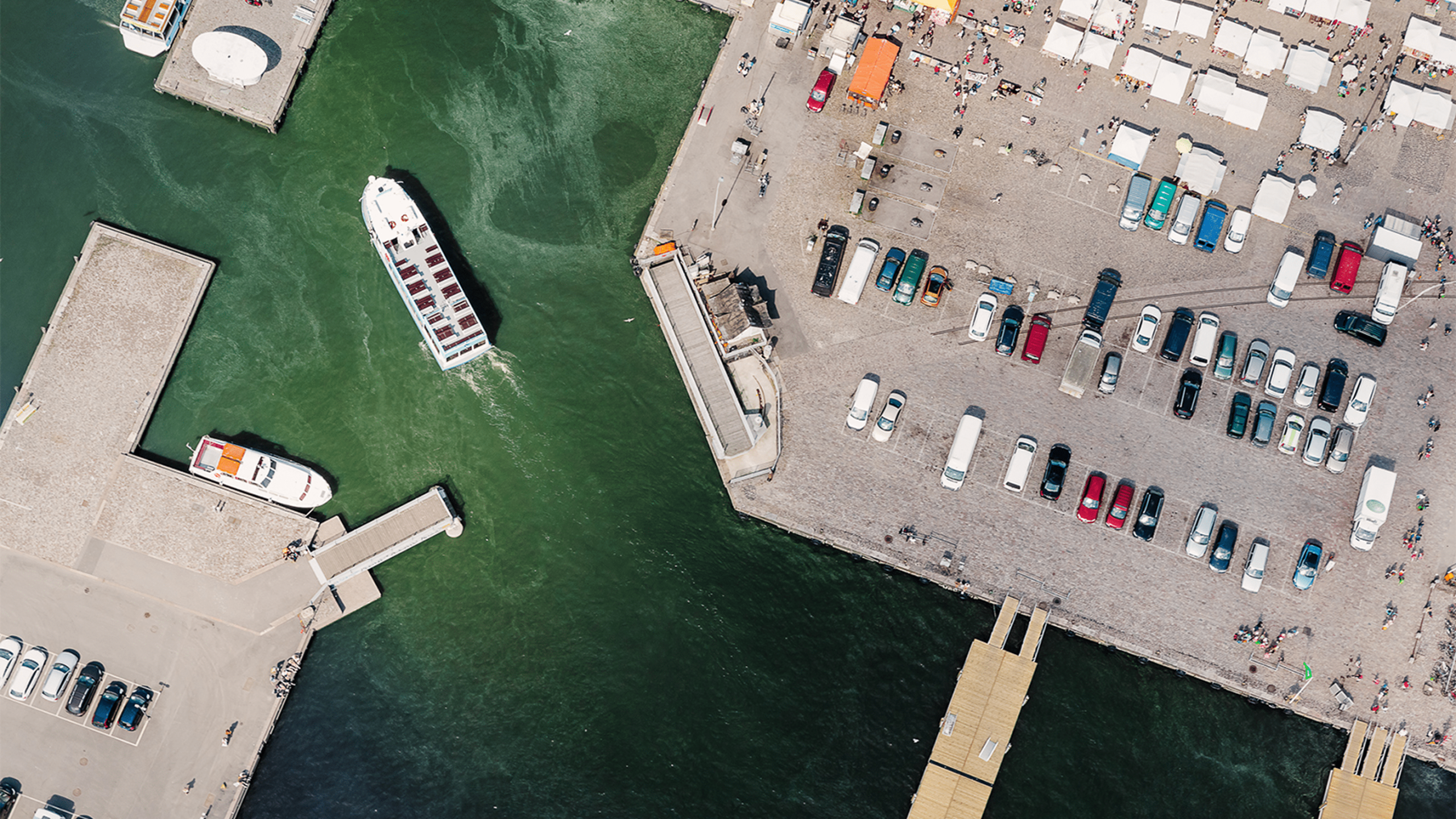 Aerial view of Helsinki's Market Square harbour with ferries, parked cars, market stalls, and green algae-tinged Baltic Sea waters.