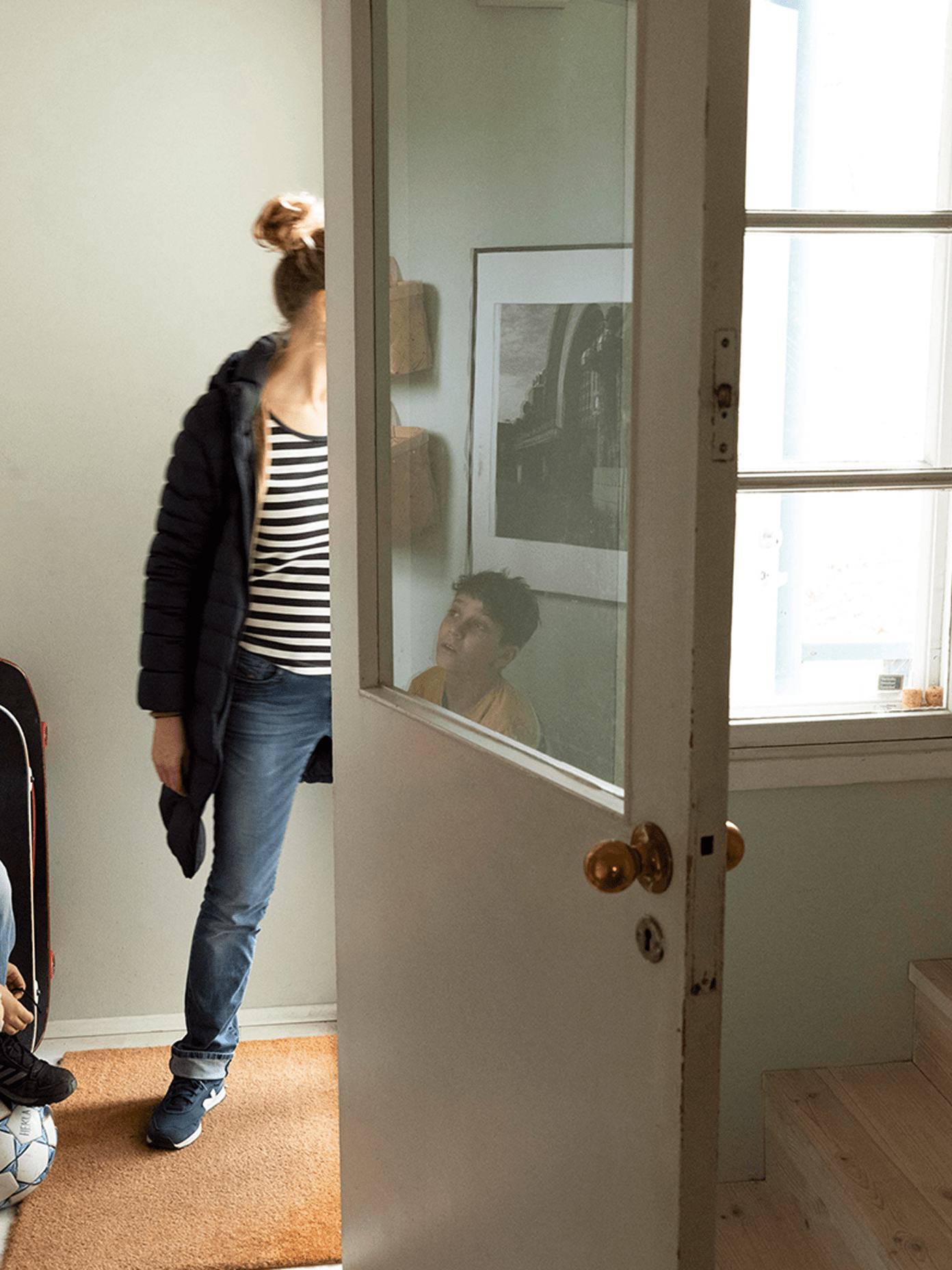 Person in striped top and puffer jacket enters a bright Helsinki apartment, while another person is reflected in a glass-paned door.