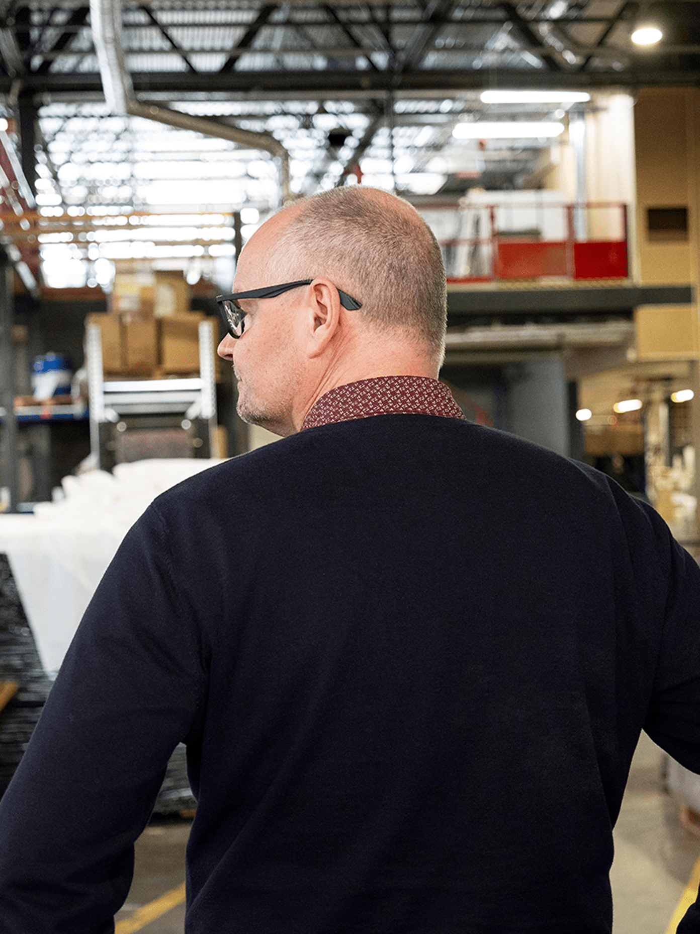 A man in a navy sweater and glasses surveys a industrial warehouse floor at Pro's manufacturing facility.