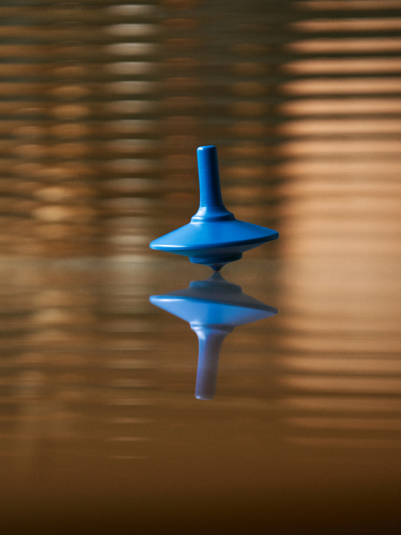 Blue spinning top balanced on its tip on a reflective surface, with warm striped light patterns in the background.