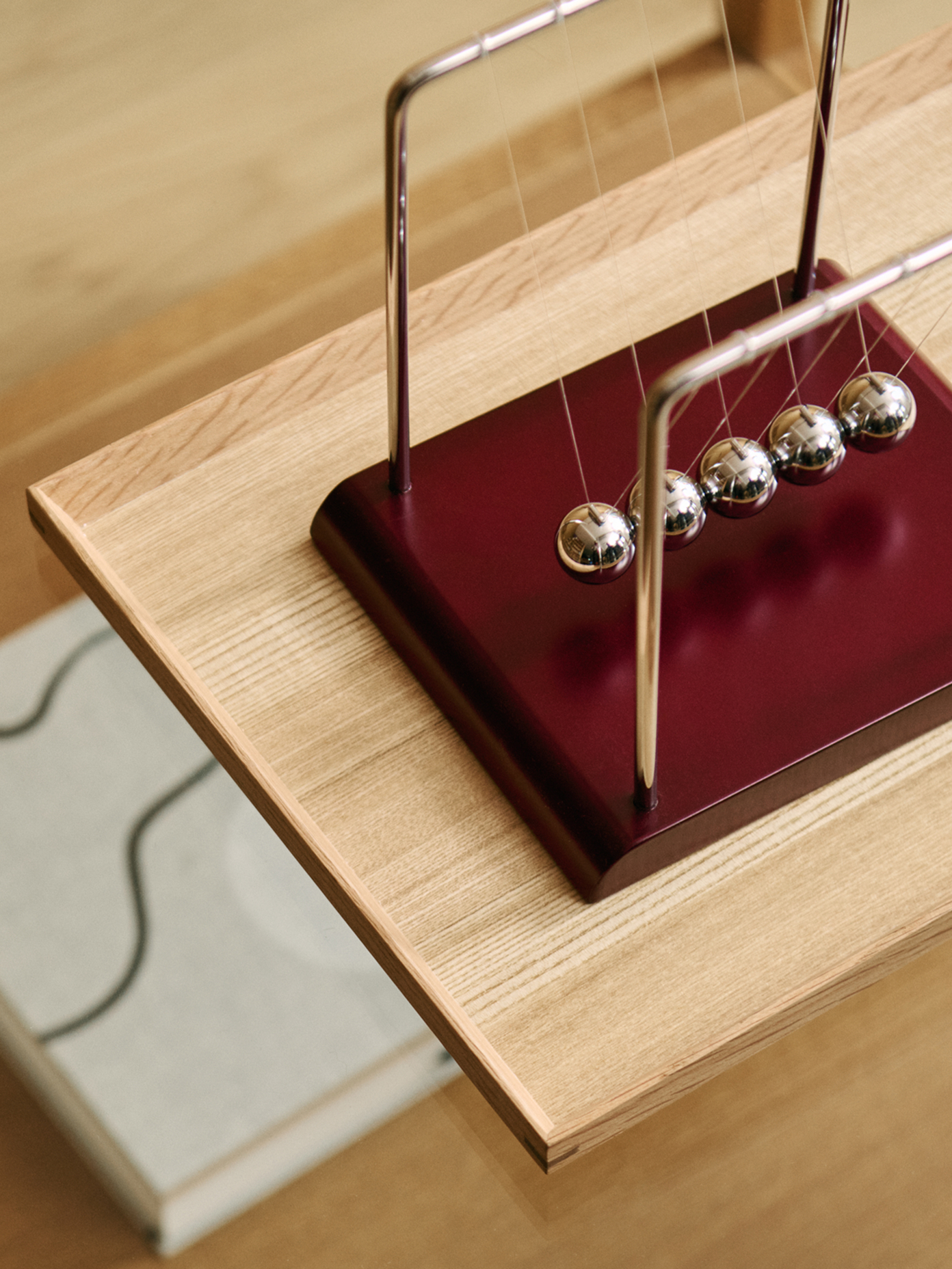 Avance Newton's cradle with chrome balls on a deep red base, placed on a light oak wooden surface.