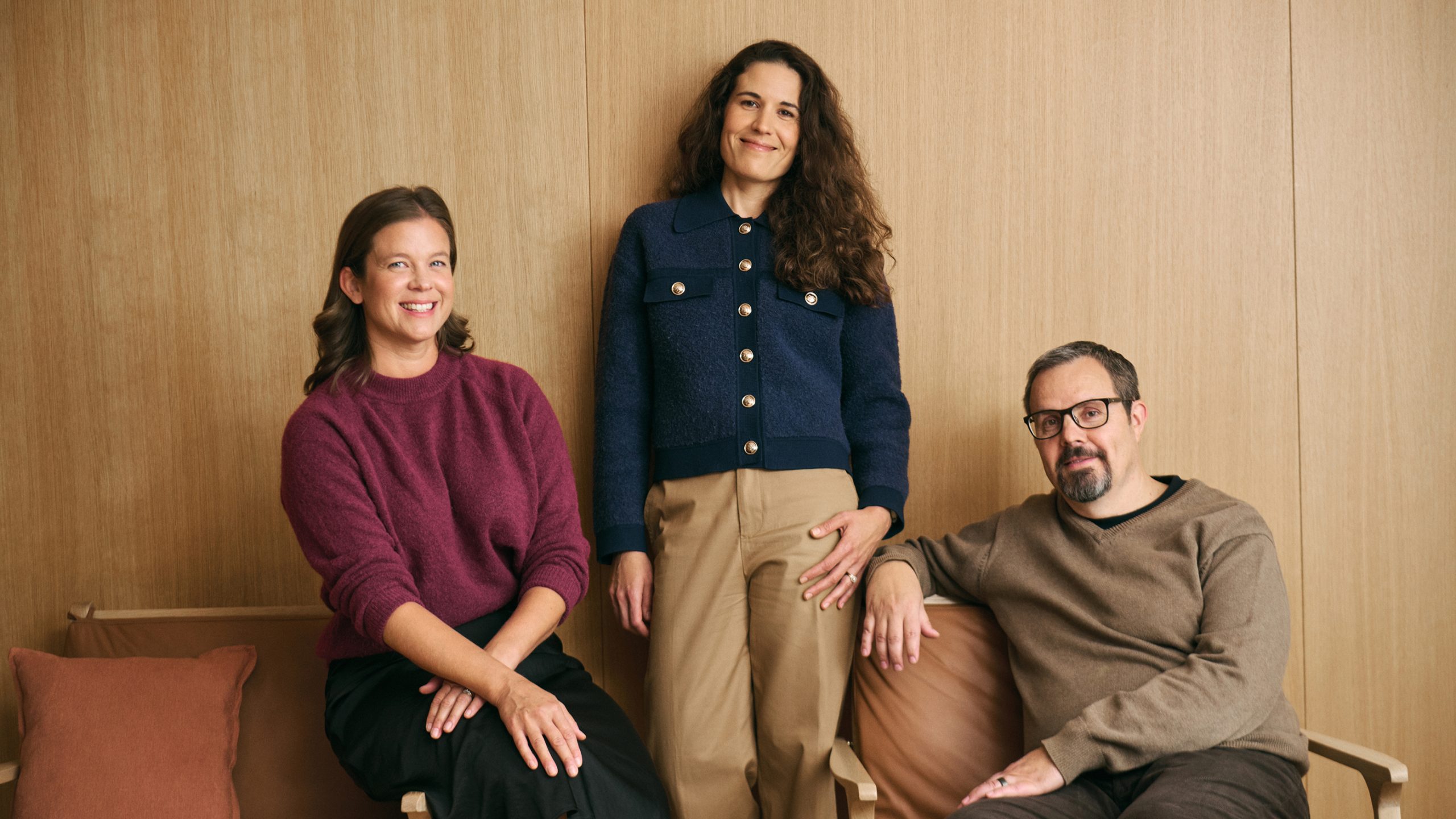 Three team members pose together against a warm wood-panelled wall in a relaxed studio setting.