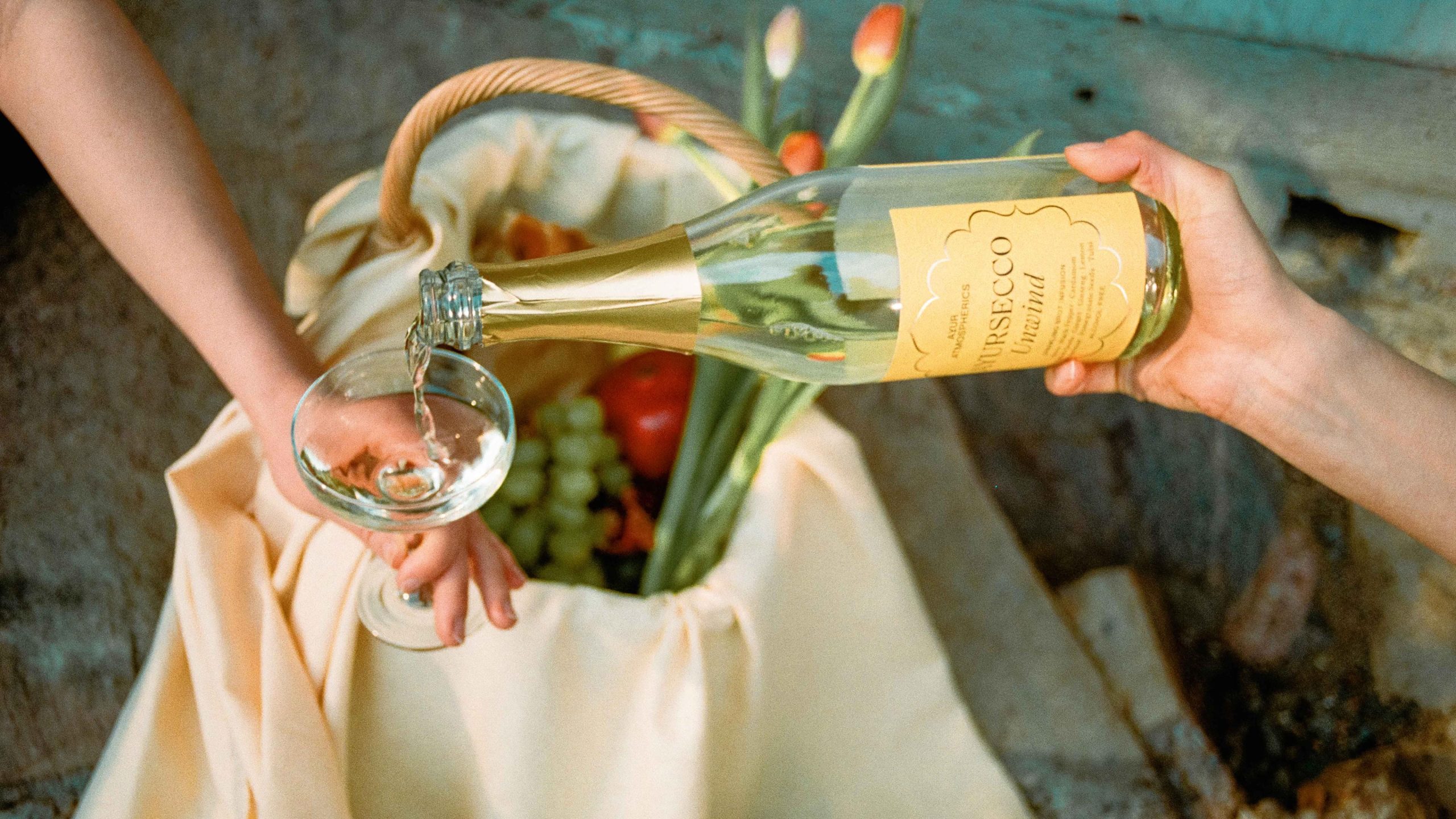 Ayur Atmospherics Ursecco Unwind sparkling bottle poured into a coupe glass over a picnic basket with fruit and tulips.