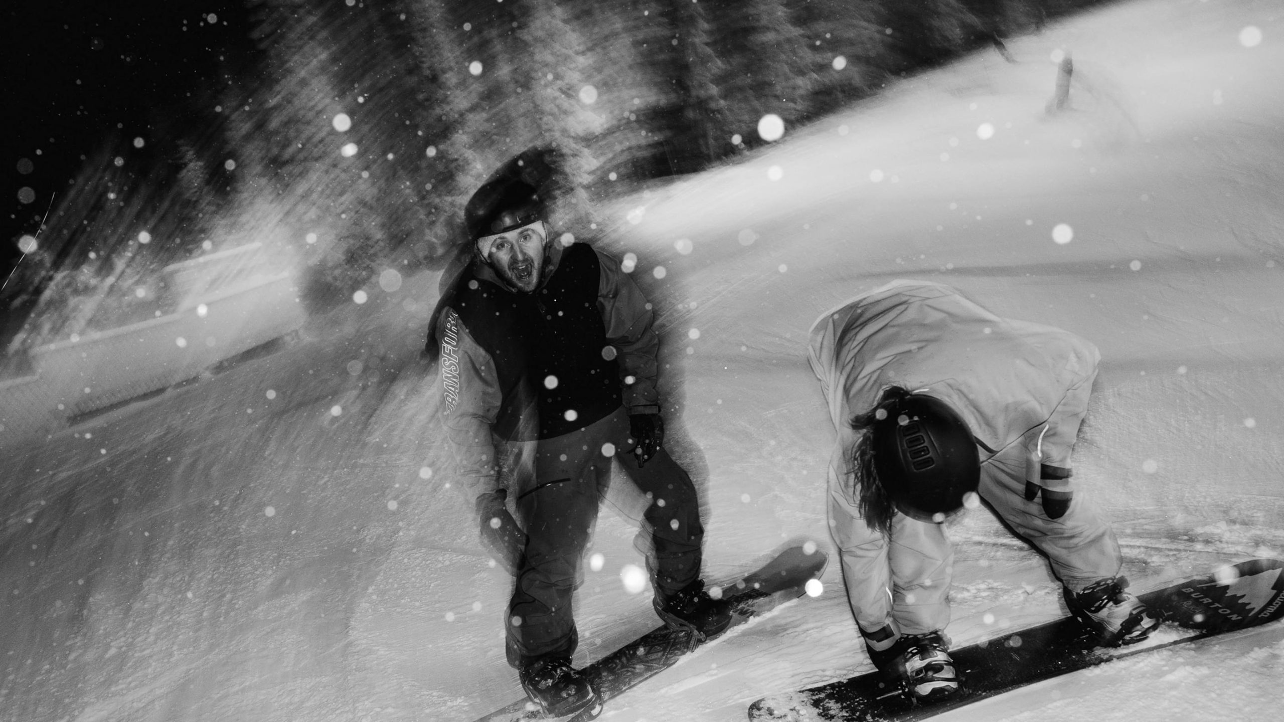 Two snowboarders carving a snowy slope at night in a blizzard, one facing camera with mouth open mid-shout.
