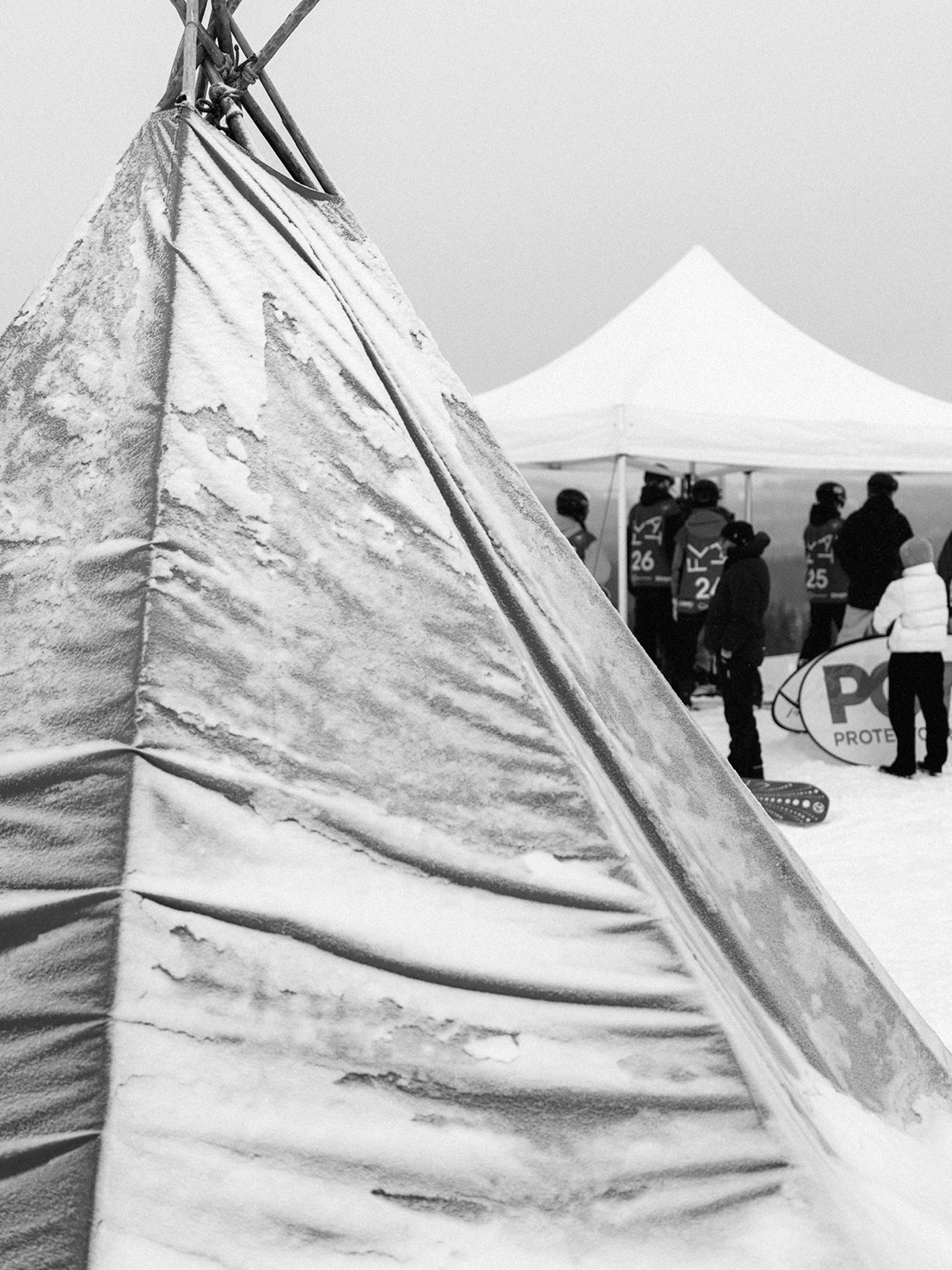 Snow-dusted tipi at a winter sports event with numbered competitors gathered under a white canopy tent.