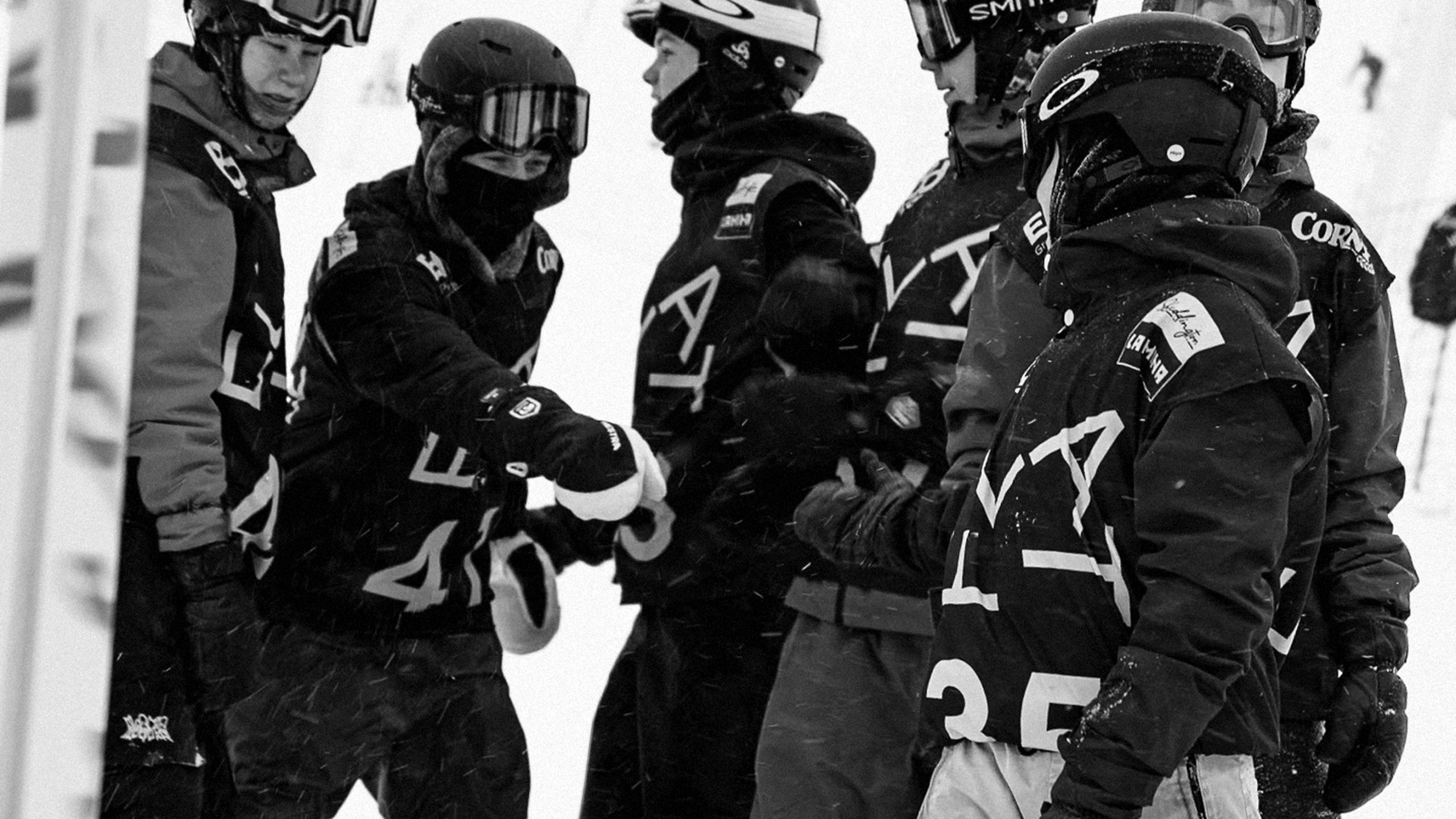 FLAT-branded skiers in matching black uniforms with race bibs fist-bump on a snowy slope during a competition.