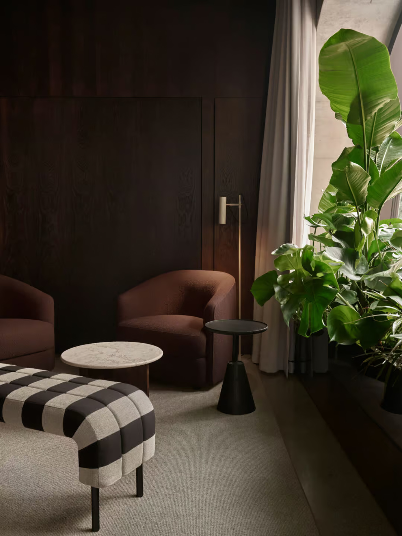 Lounge corner with checkered ottoman, mauve armchair, marble side table, brass floor lamp, and lush tropical plants by a window.
