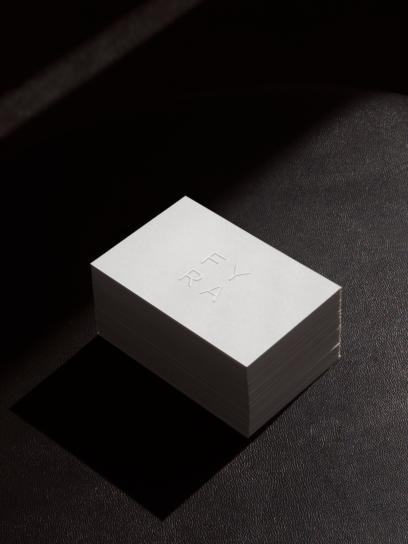 Stack of white Fyra business cards with blind embossed logo on dark leather surface, dramatic side lighting.