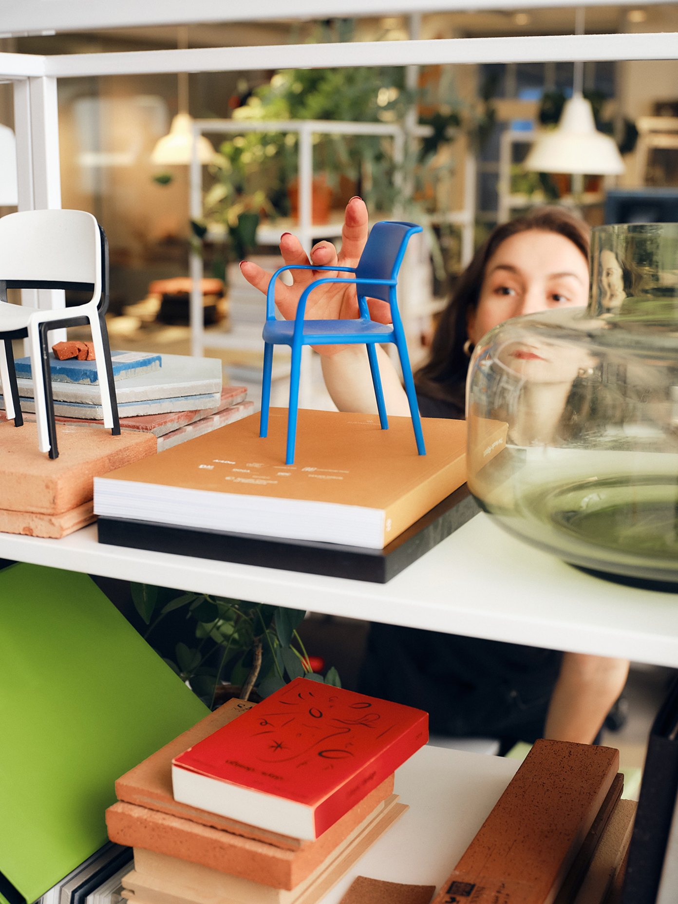 A woman peers through a shelf holding a miniature blue chair model, surrounded by design books and material samples.
