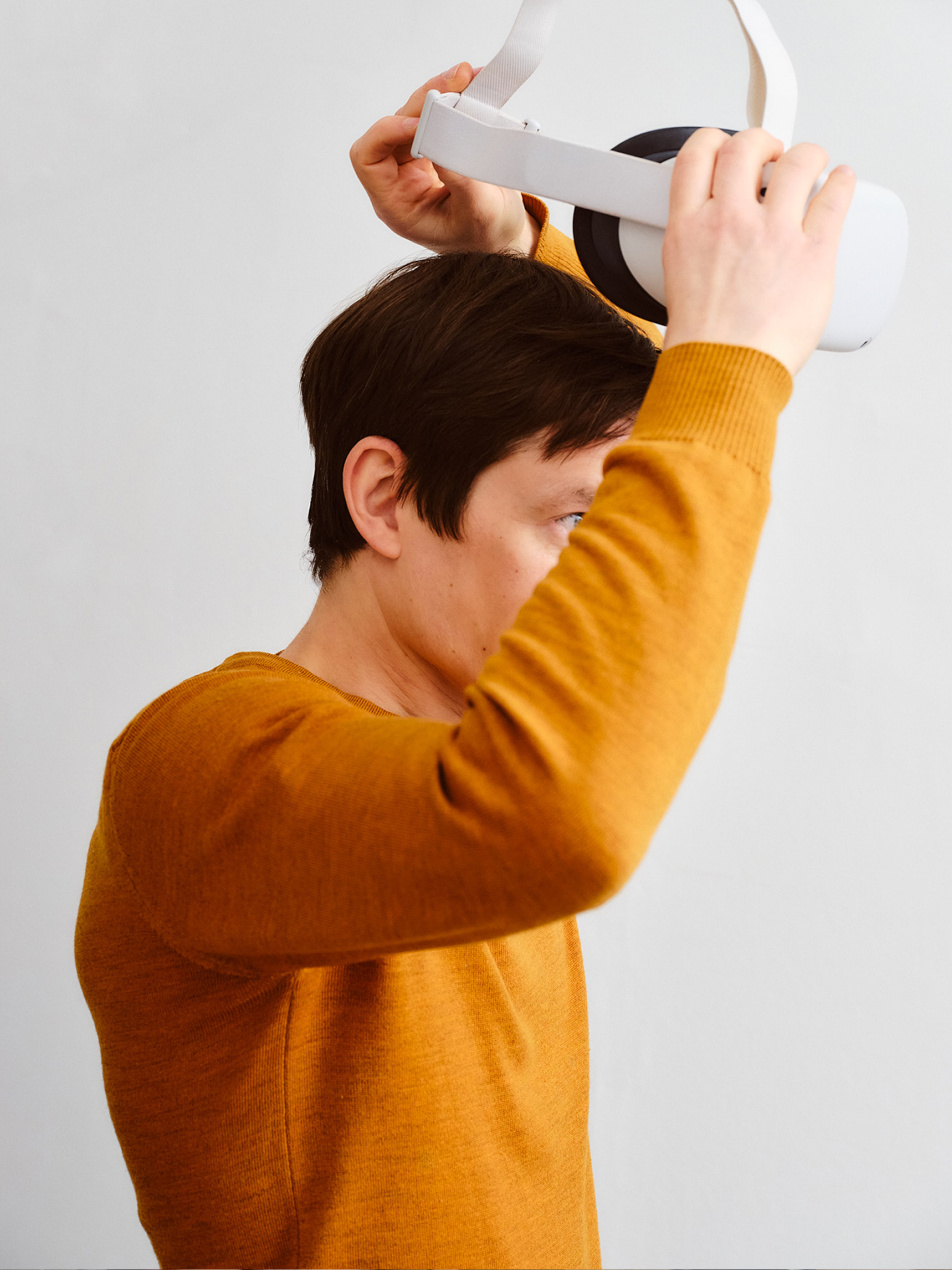 Person in mustard yellow sweater putting on a white VR headset against a light grey background.