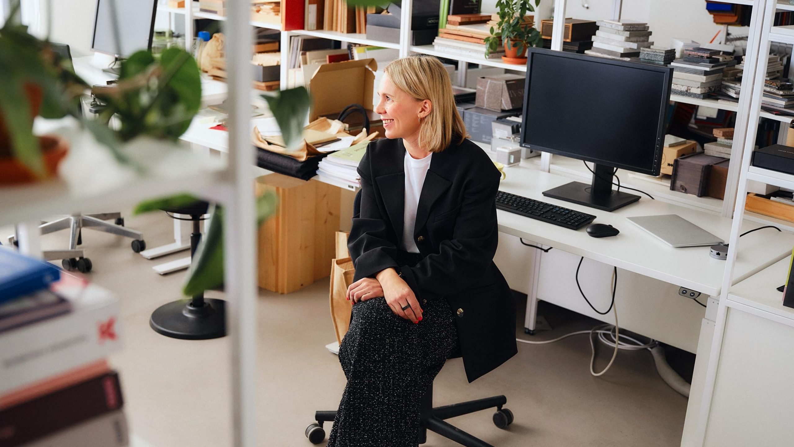 A smiling blonde woman in a black blazer sits at a design studio desk surrounded by shelves of books and material samples.