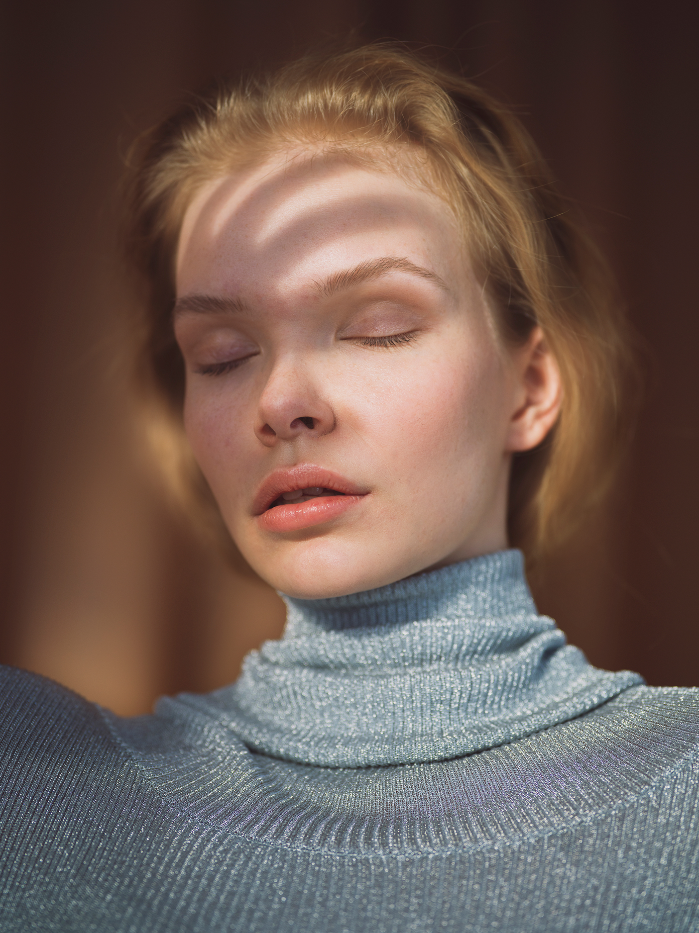 Young woman with closed eyes and short blonde hair wearing a silver-blue turtleneck sweater.