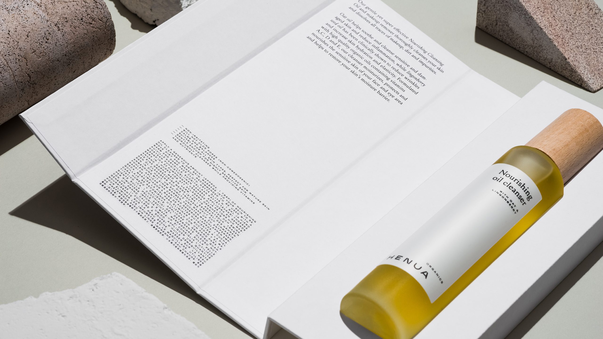Henua Organics nourishing oil cleanser bottle with wooden cap resting on open product booklet, styled with stone props.