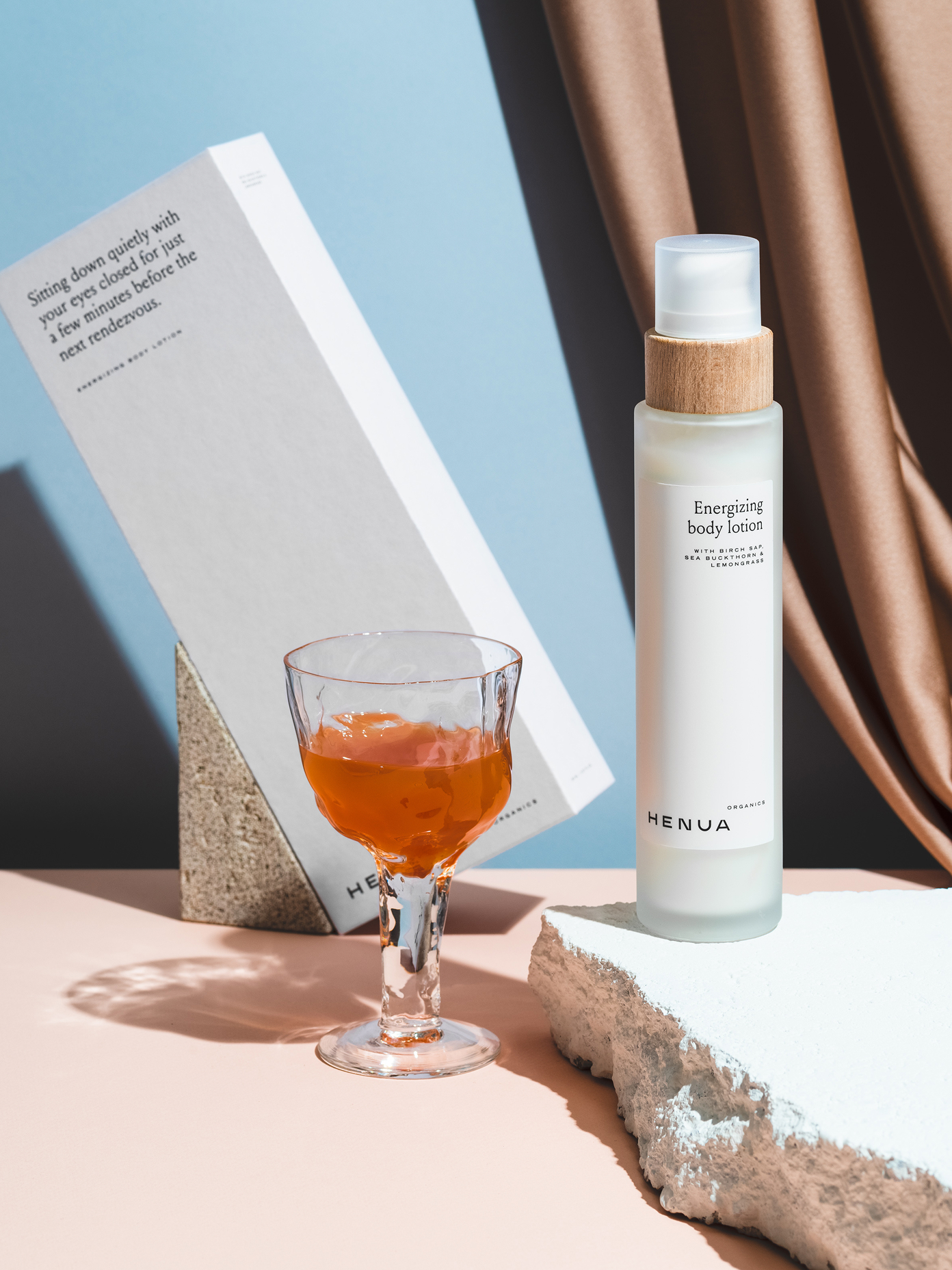 Henua Organics Energizing Body Lotion bottle with wooden cap beside a cocktail glass and branded box on a pink surface.