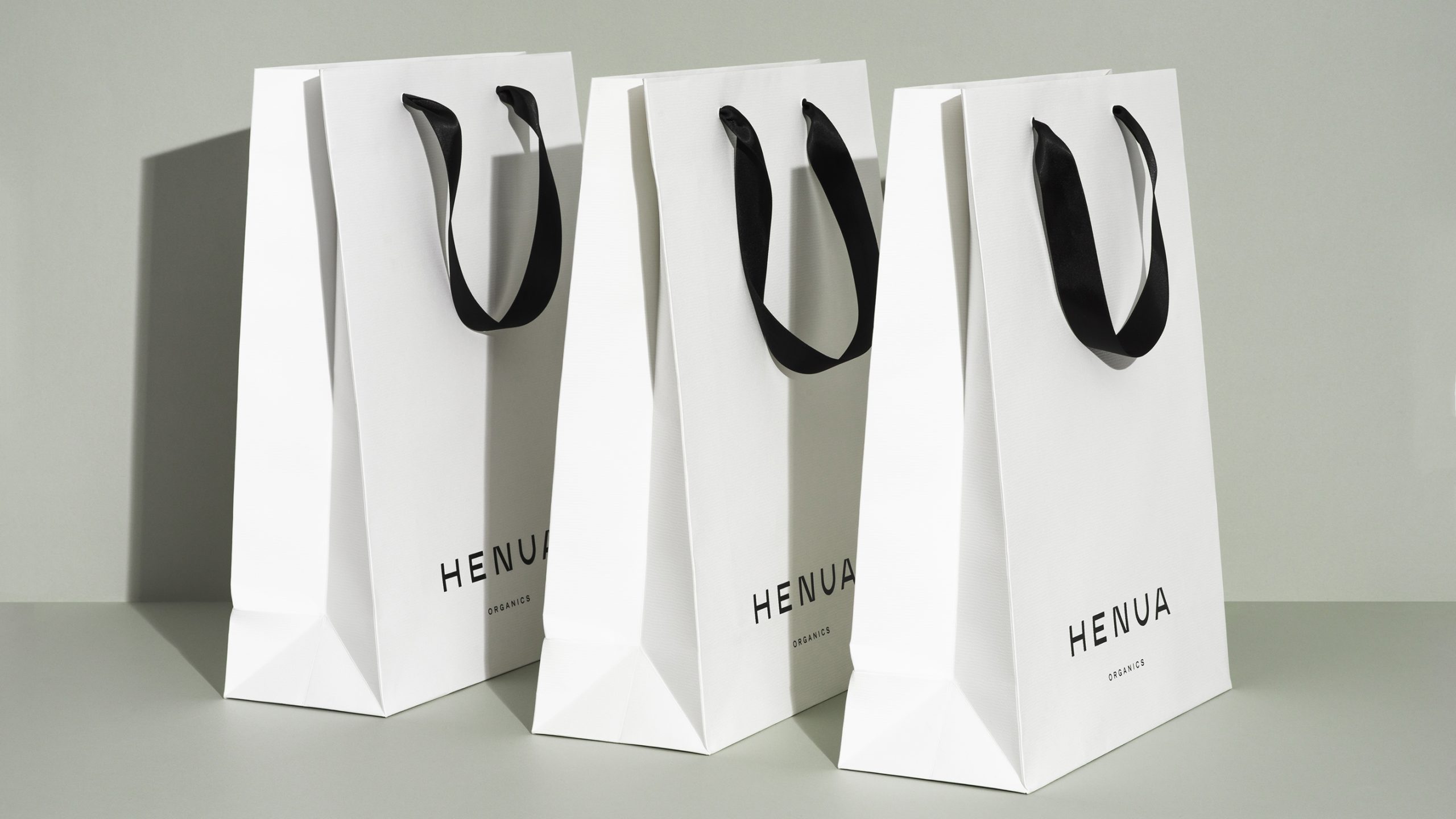 Three white Henua Organics paper shopping bags with black ribbon handles arranged on a grey surface.