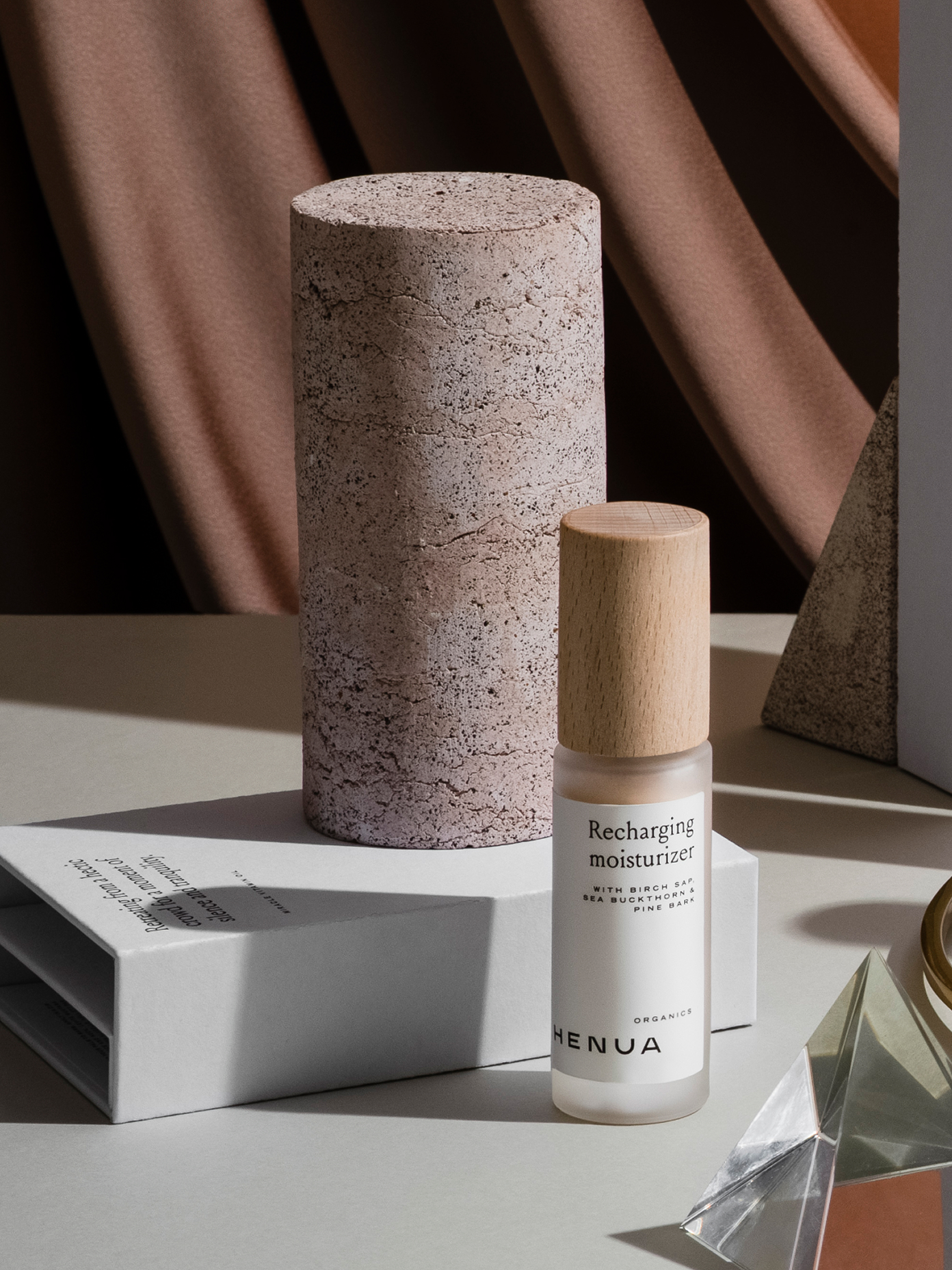 Henua Organics Recharging Moisturizer bottle with wooden cap beside a stone cylinder and branded box.