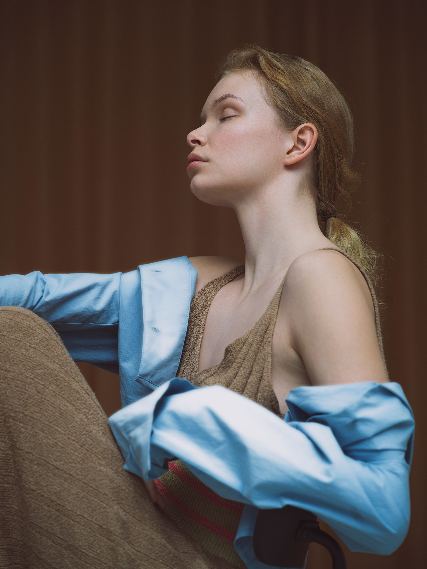 Model in a light blue oversized shirt and beige knit top, eyes closed, seated against a warm brown background for Henua.