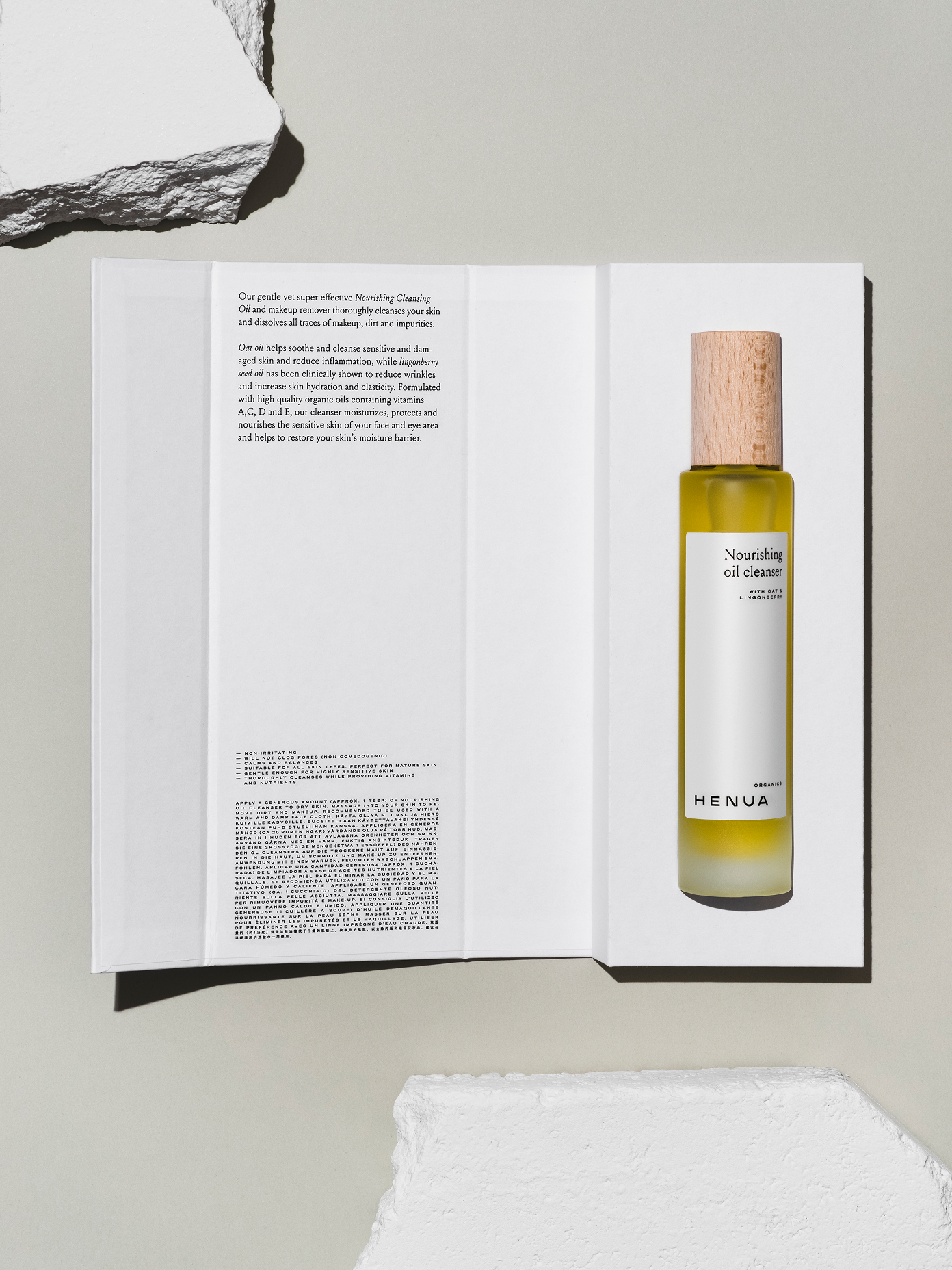 Henua Organics Nourishing Oil Cleanser bottle with wood cap resting on open product booklet, styled with raw stone props.