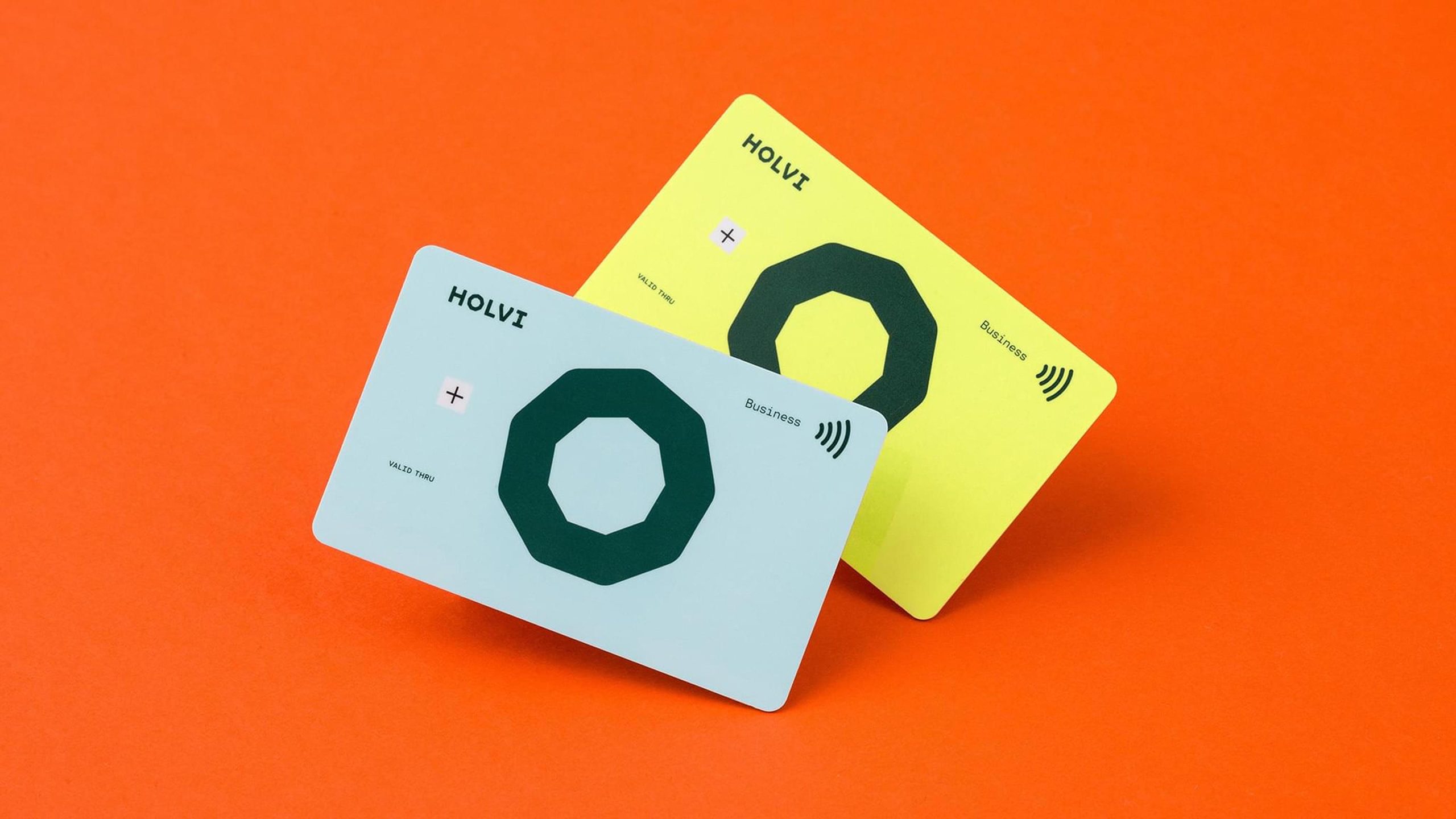 Two Holvi Business debit cards in pale blue and neon yellow, featuring a dark green octagon logo, displayed on an orange background.