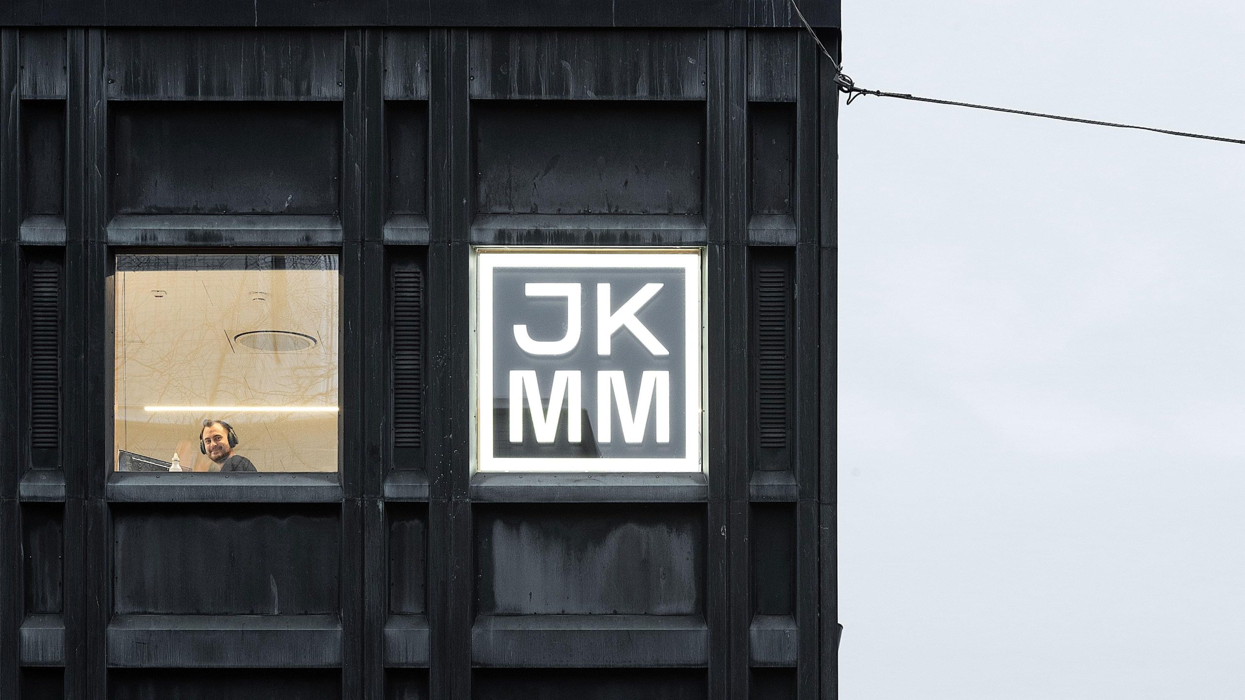 JKMM Architects illuminated logo sign on a dark timber-clad building facade.