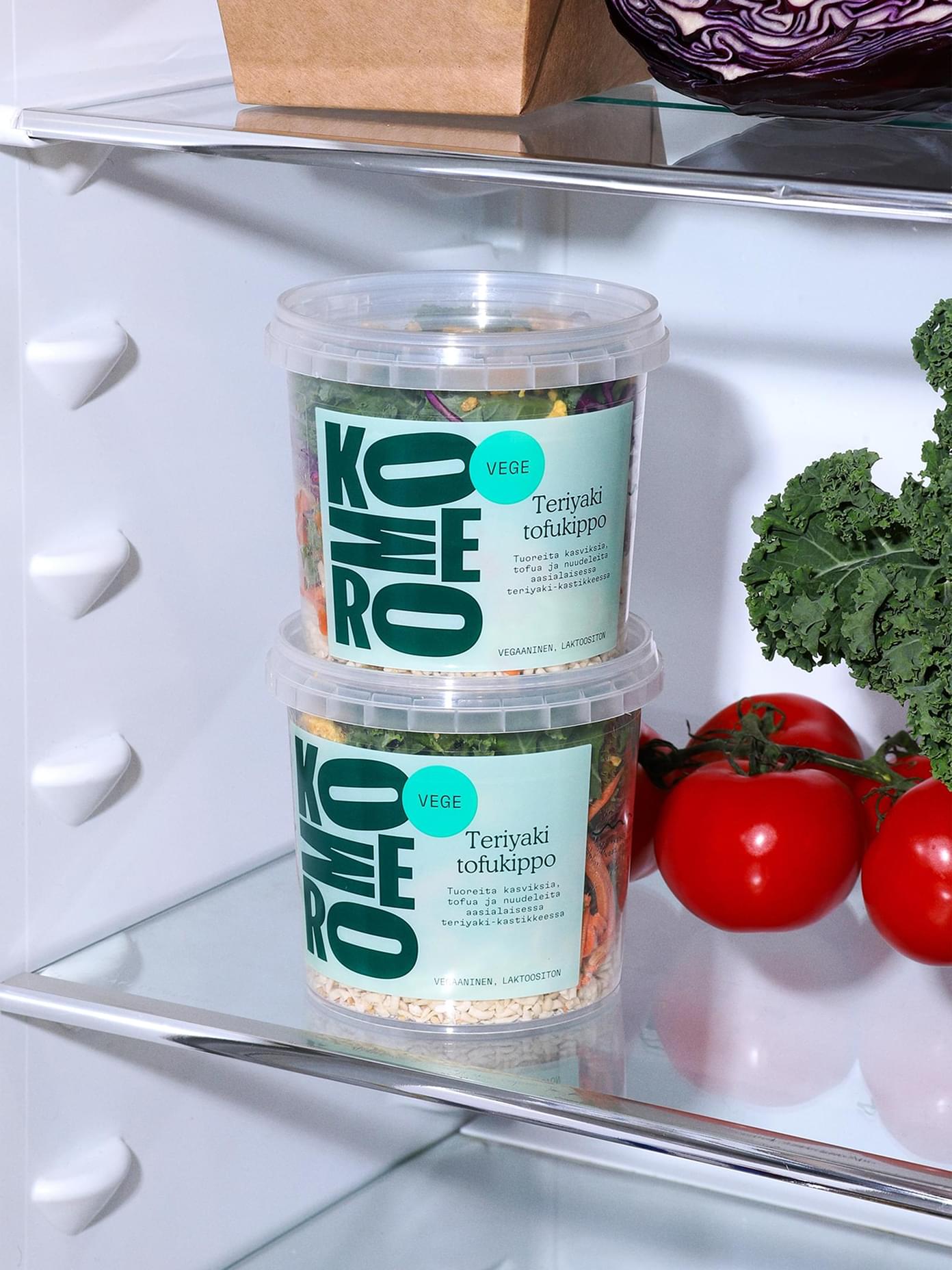 Two Komero Teriyaki tofukippo vege meal containers stacked on a refrigerator shelf beside kale and cherry tomatoes.
