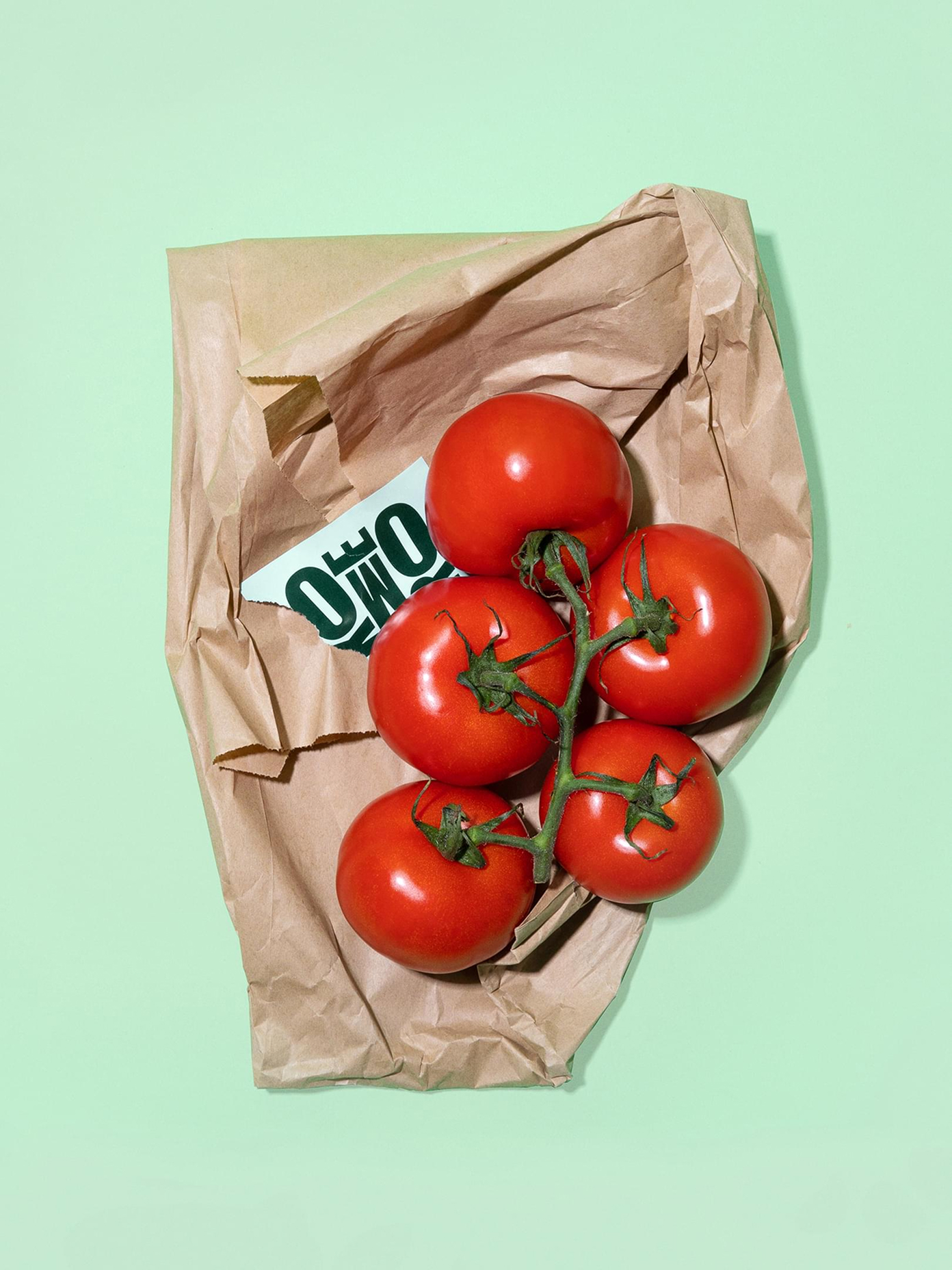 Vine tomatoes spilling from a torn kraft paper bag with a Komero label, styled on a mint green background.