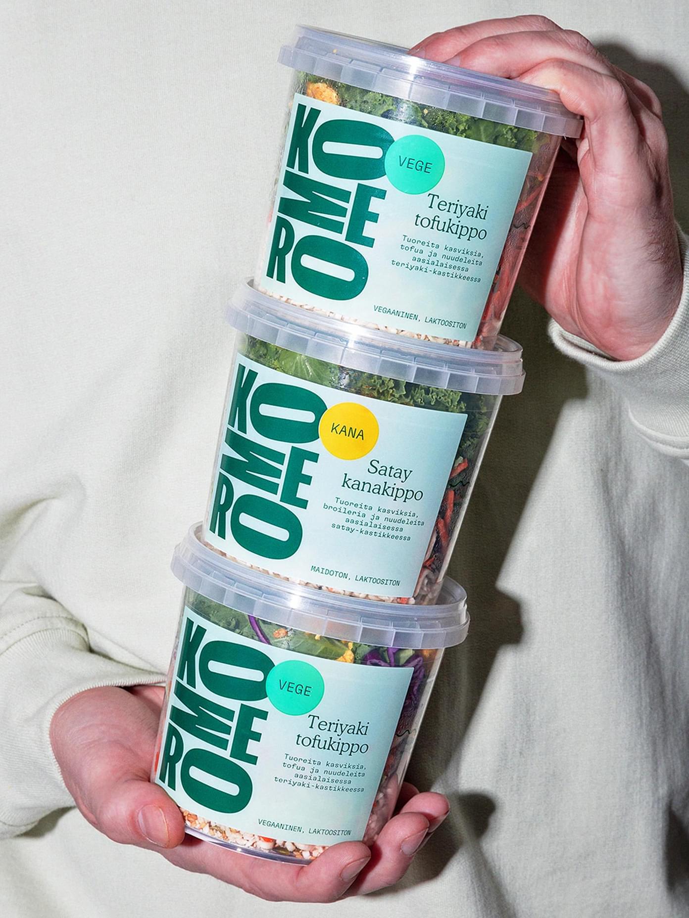 Hands holding three stacked Komero food containers with Finnish labels for Teriyaki tofukippo and Satay kanakippo.