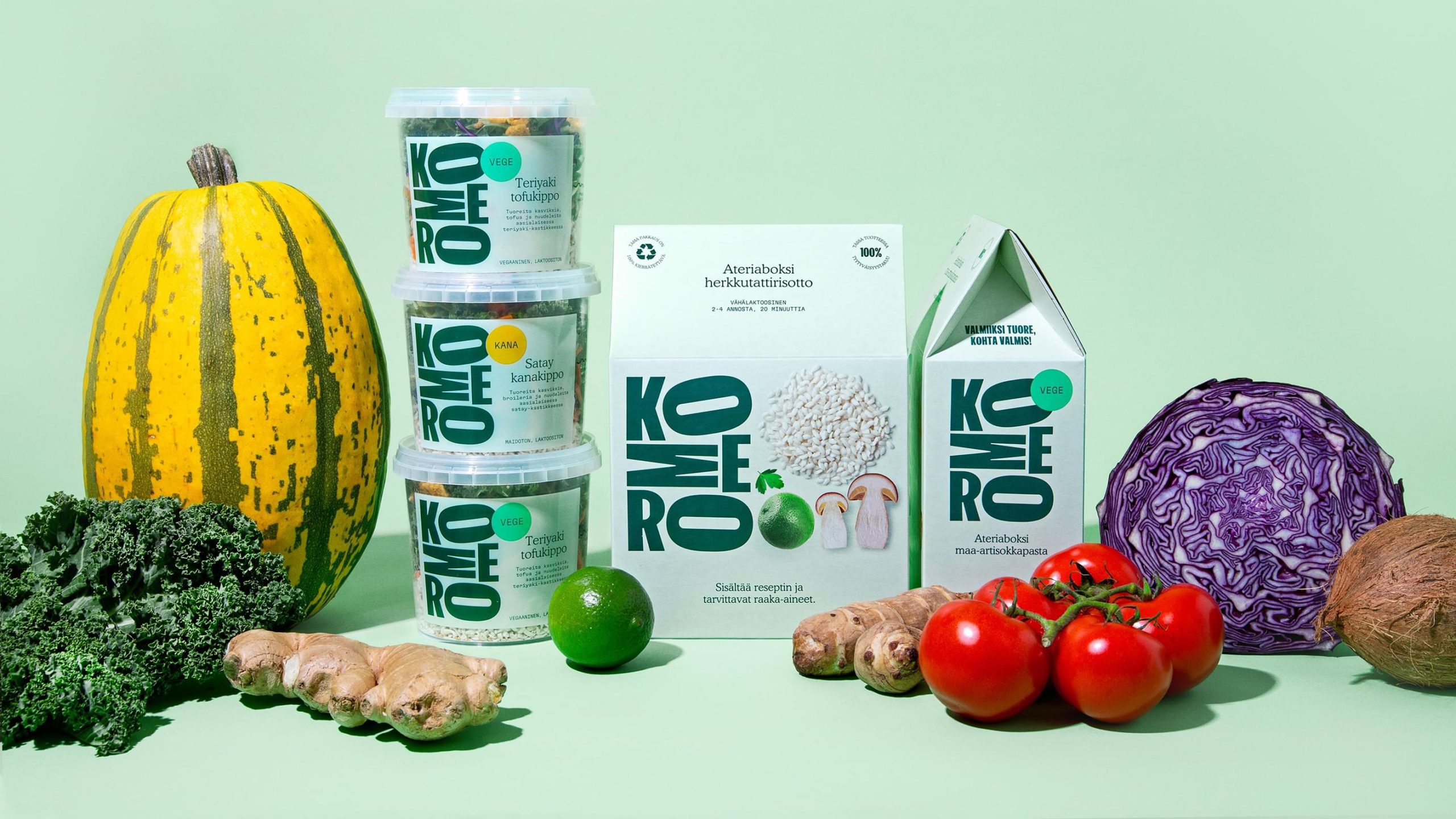 Komero meal kit packaging surrounded by fresh vegetables including squash, kale, tomatoes, ginger, lime, and red cabbage.