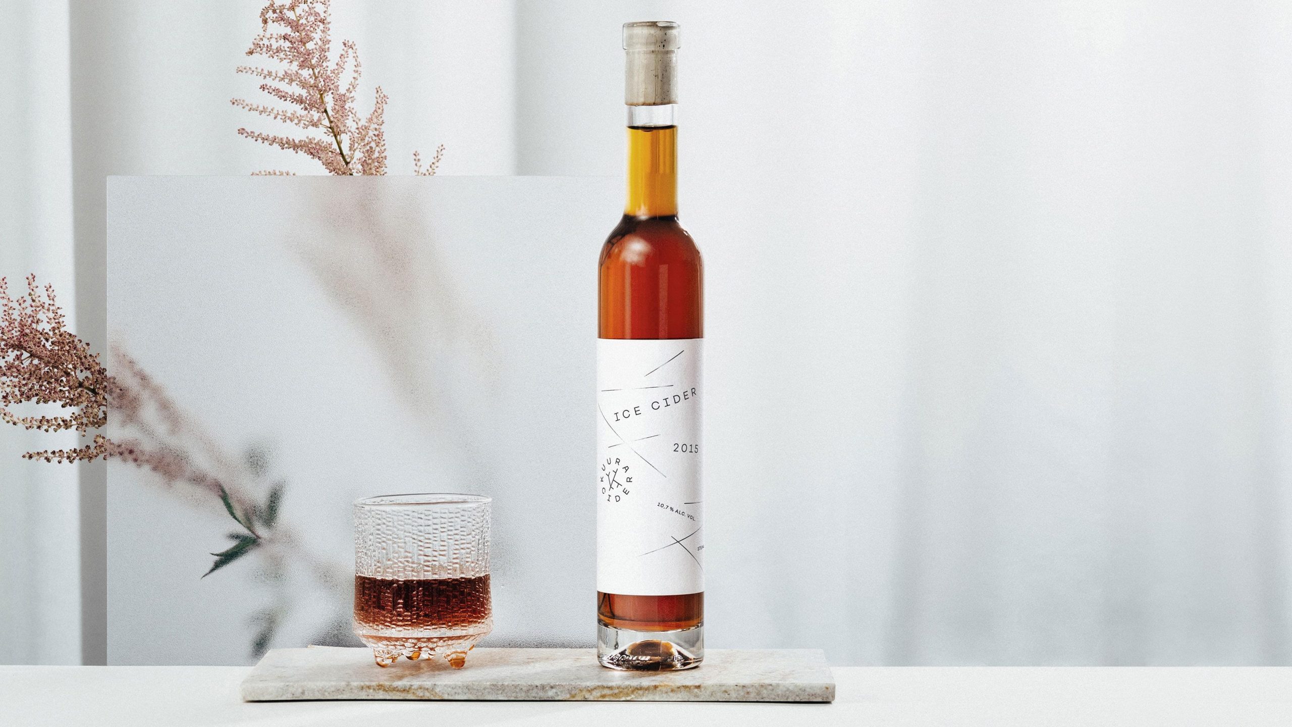 Kuura Ice Cider 2015 bottle with minimalist white label beside a textured glass on a marble slab, styled with dried botanicals.