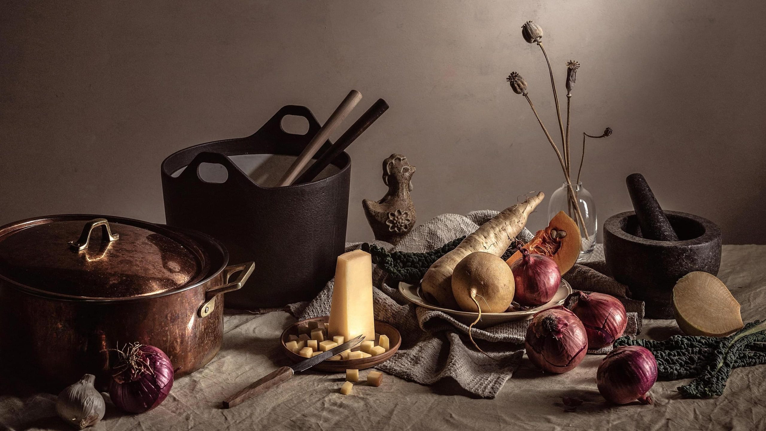 Kuusamon Juusto cheese block with onions, root vegetables, copper pot, mortar, and dried florals in a moody still life.