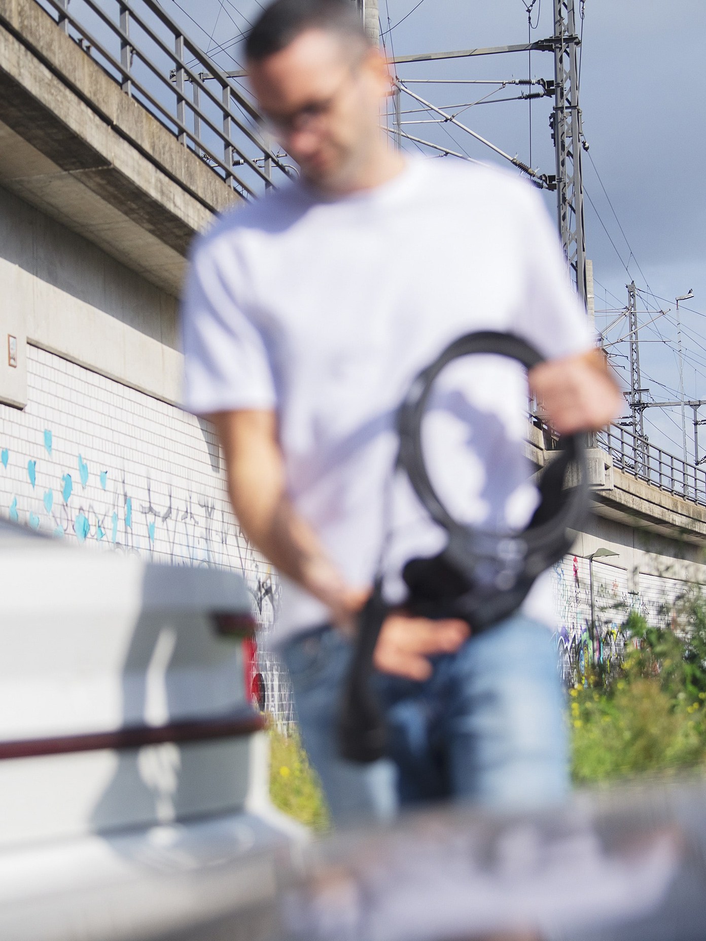 Person in white t-shirt holding camera equipment near graffiti wall and railway bridge, shot with shallow depth of field.
