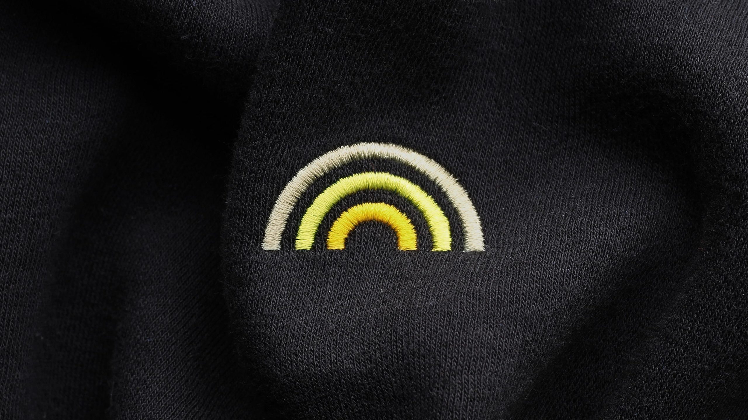 Embroidered Plugsurfing rainbow logo in yellow and white on a black fabric garment.