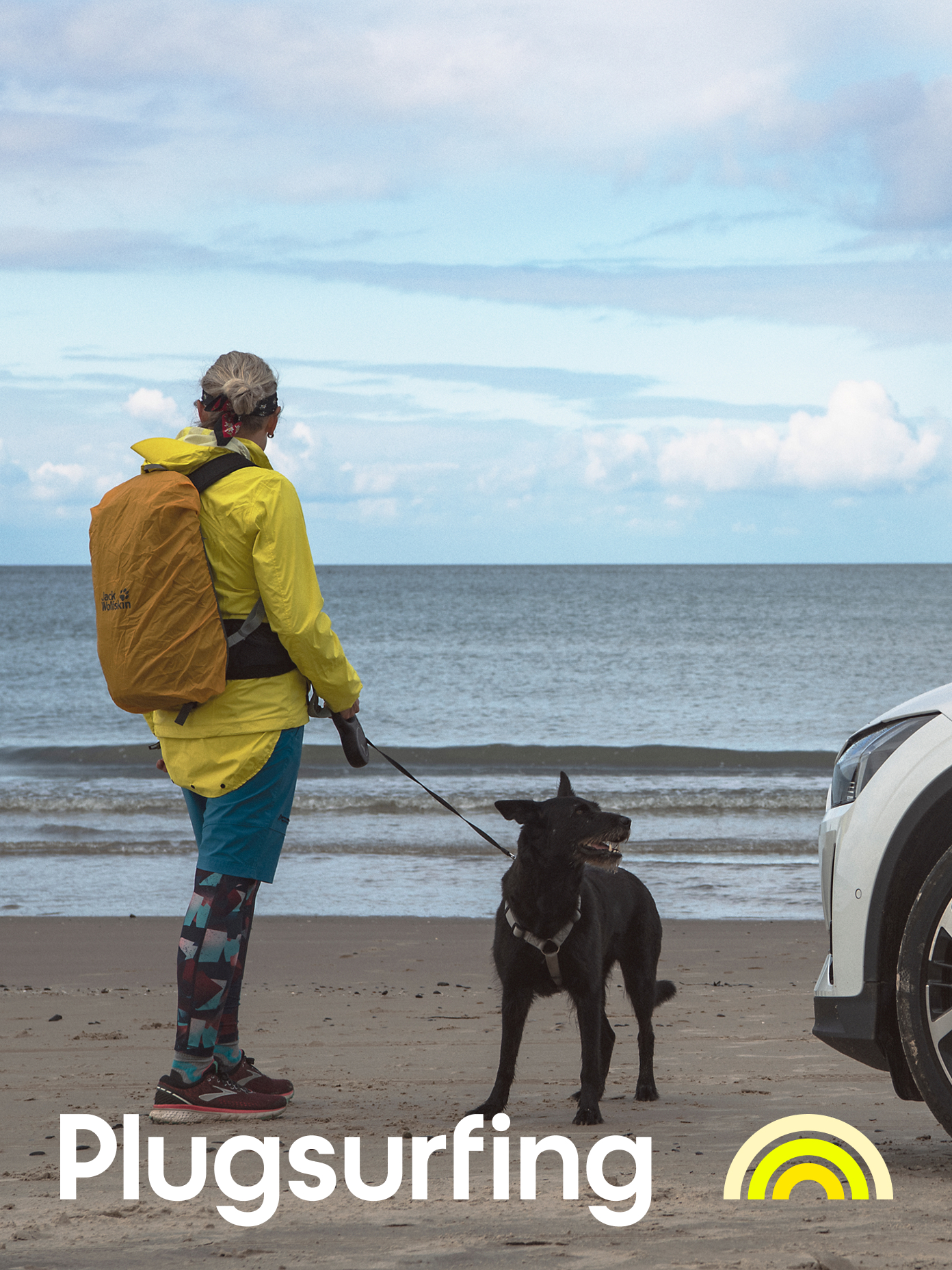 Person in yellow jacket with black dog on leash stands by white EV on a sandy beach, Plugsurfing branding visible.