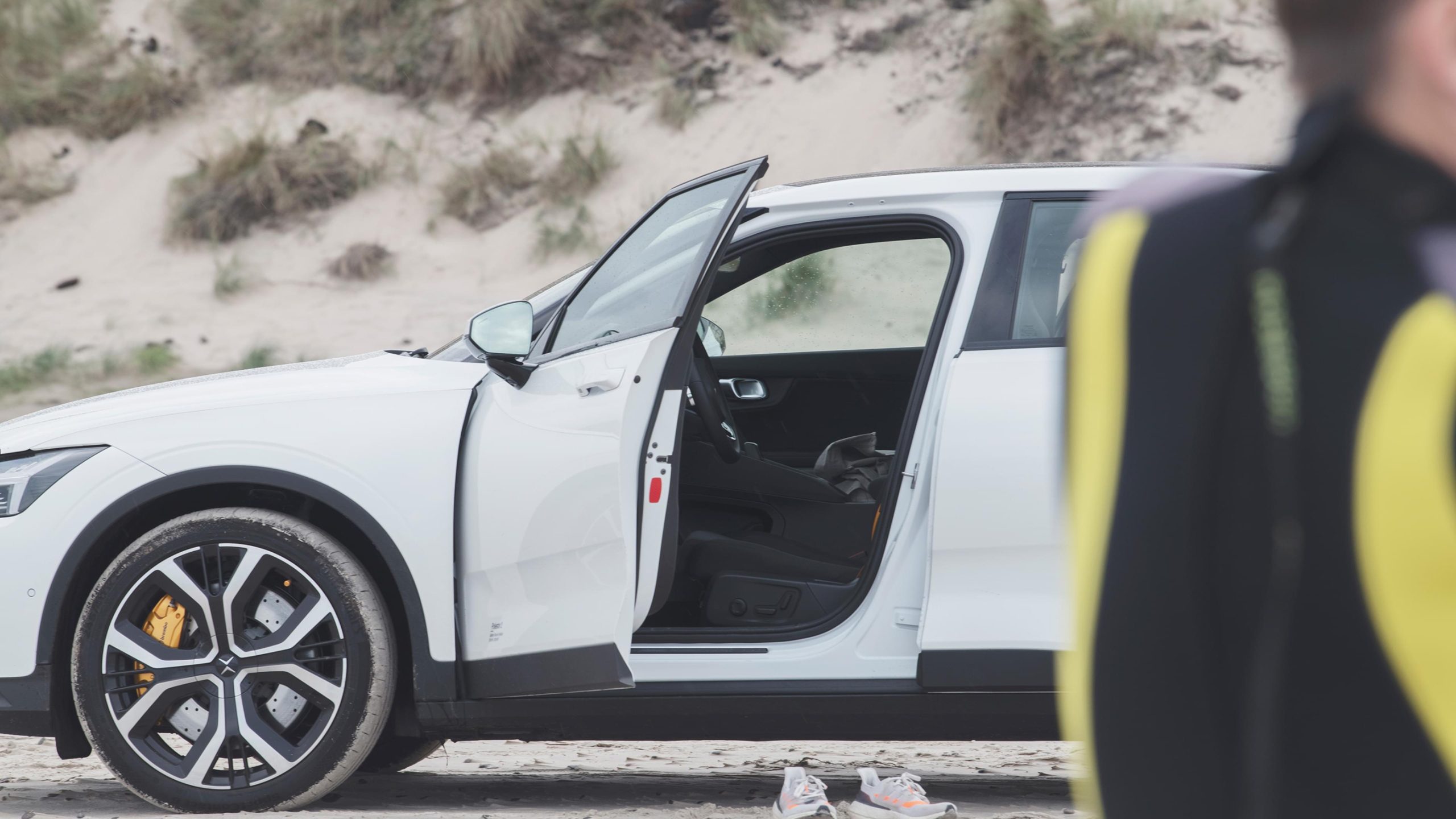 White electric car with open door parked on a sandy beach, yellow brake calipers visible on the alloy wheel.