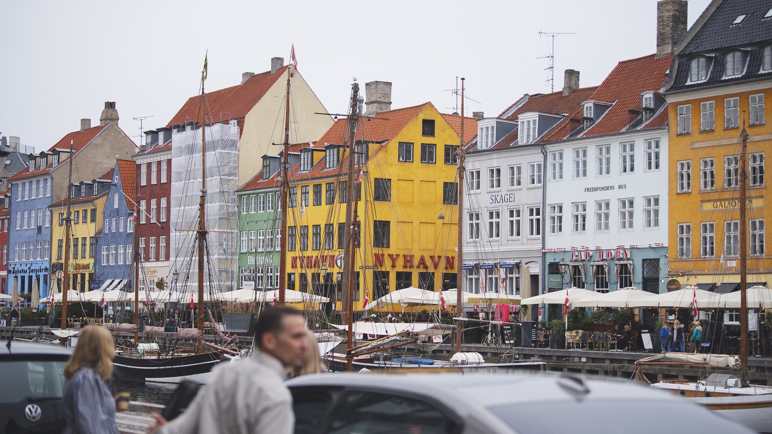 Waterfront in Copenhagen with colourful historic buildings, sailing boats, and outdoor restaurant terraces.