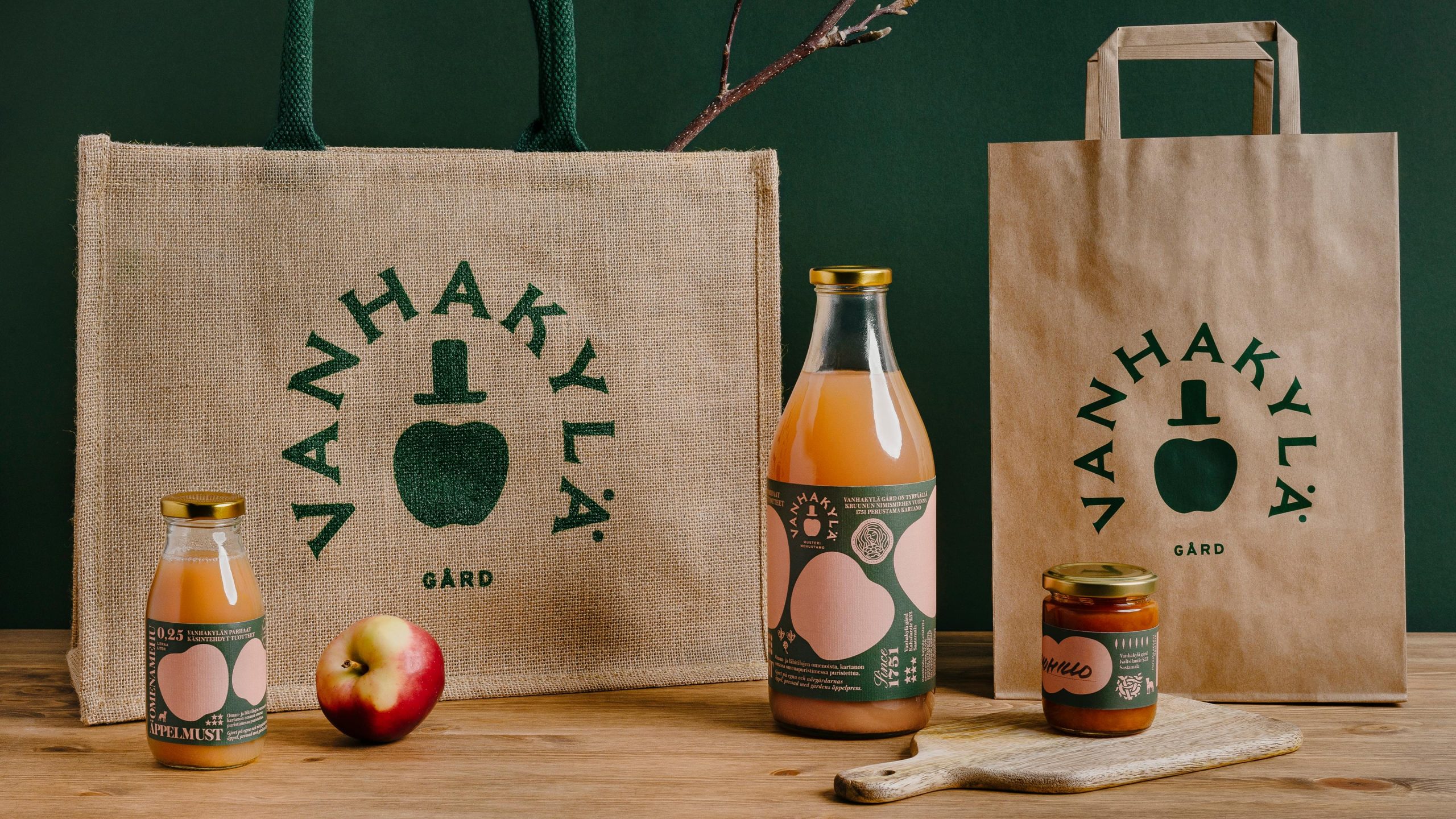 Vanhakylä Gård branded packaging including jute bag, paper bag, juice bottle, small jar, and apple on a wooden surface.