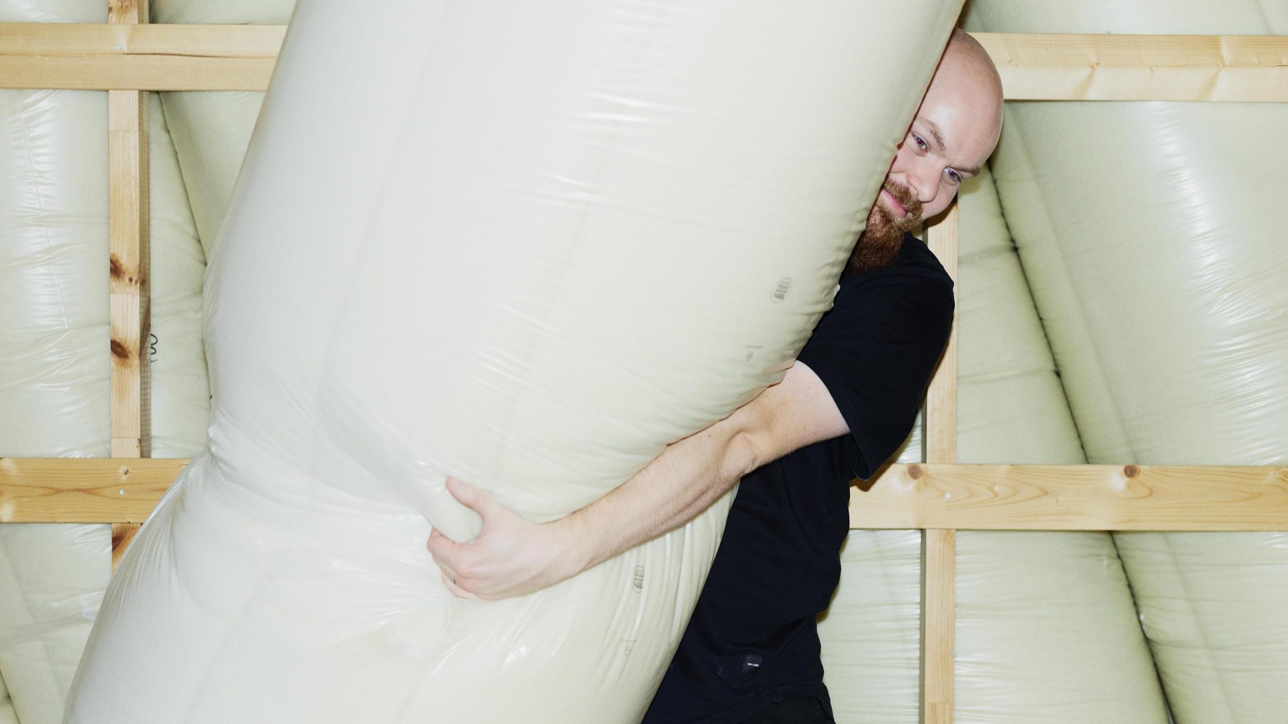 A man in black hugs an oversized cream inflatable form among stacked bulbous shapes in a wooden-framed space.
