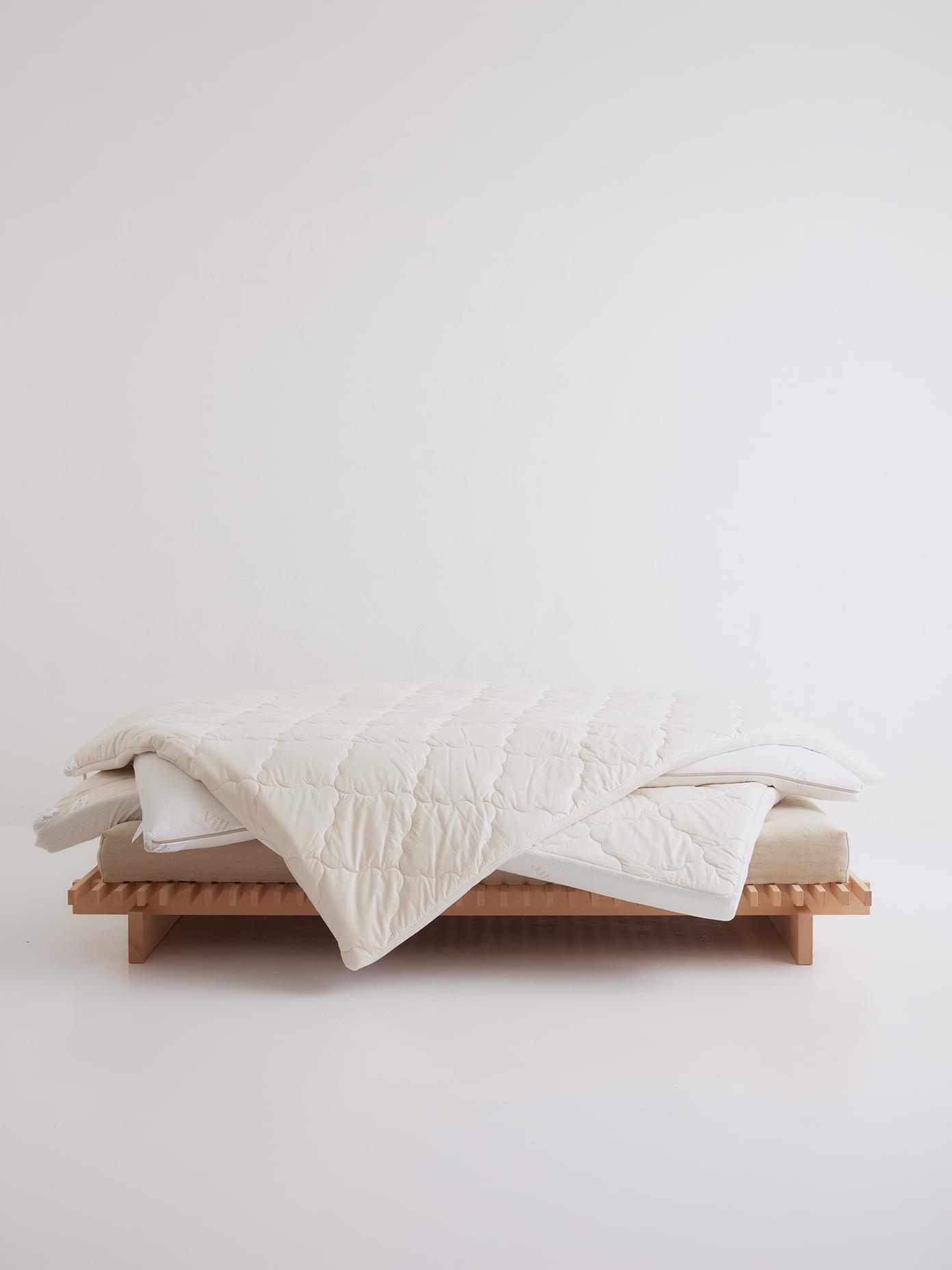 Villa & Peite quilted white comforter draped over pillows on a low natural wood slatted bed frame against a white background.