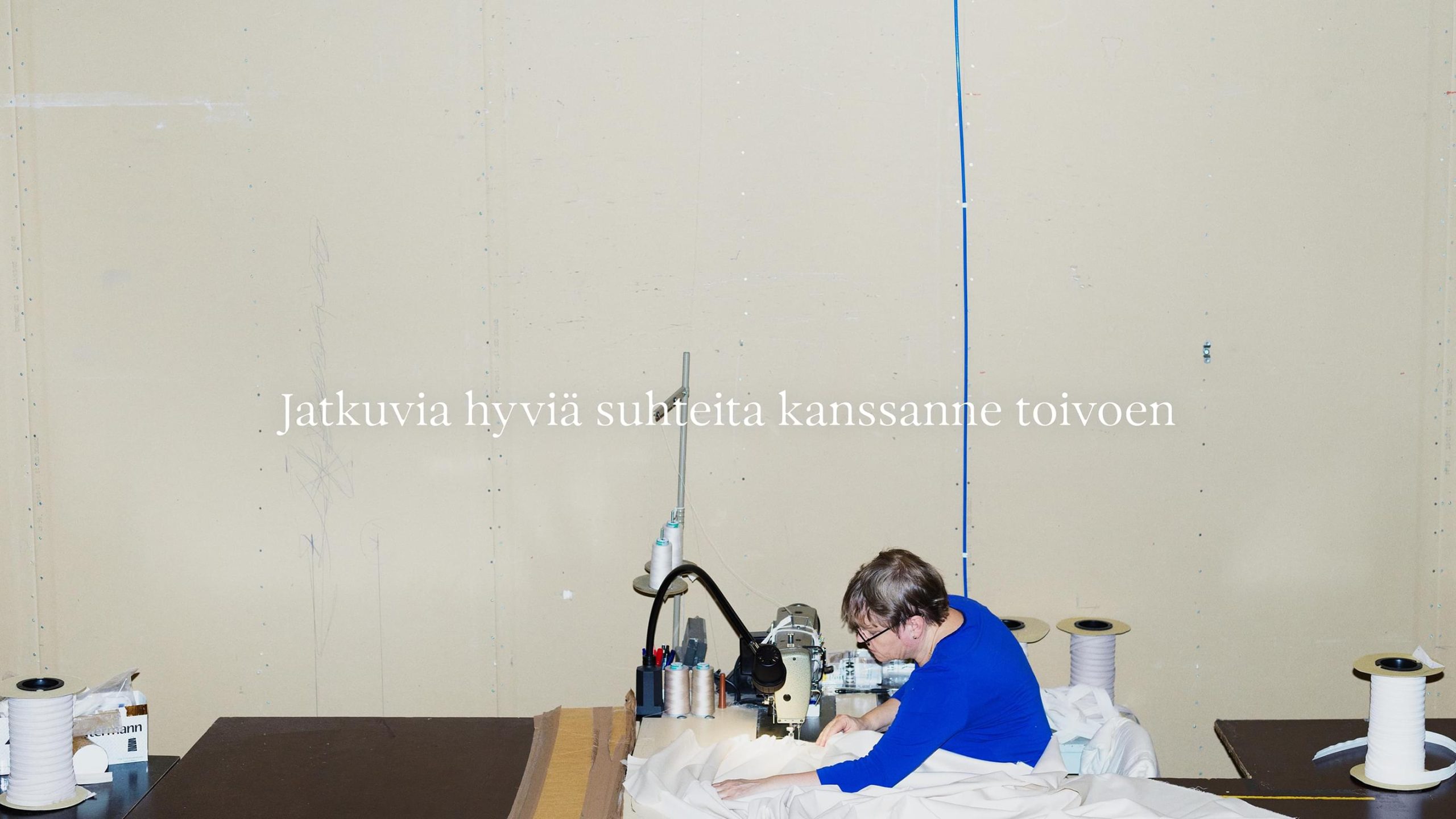 Villa & Peite seamstress in blue top working at a sewing machine surrounded by white fabric and thread spools in a workshop.